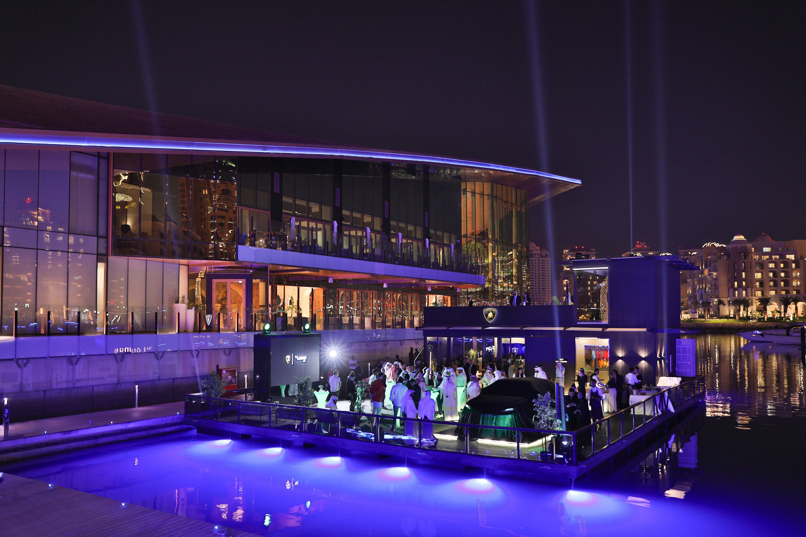 Night shot of the lamborghini lounge doha illuminated in purple and blue lighting with guests attending the opening event and a green urus on display