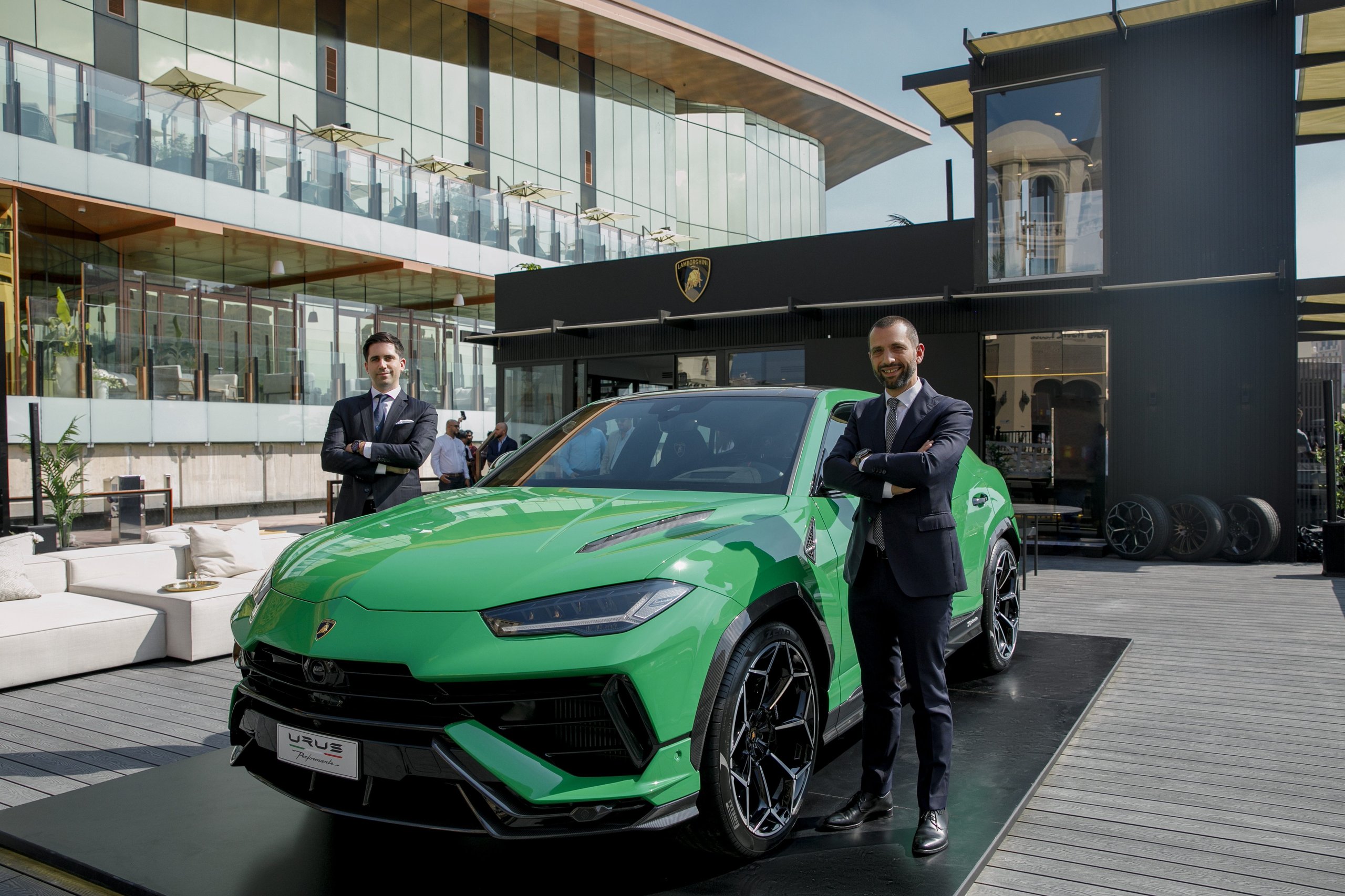 Two executives in suits standing beside a green lamborghini urus performante at the doha lounge opening with the modern glass structure in the background