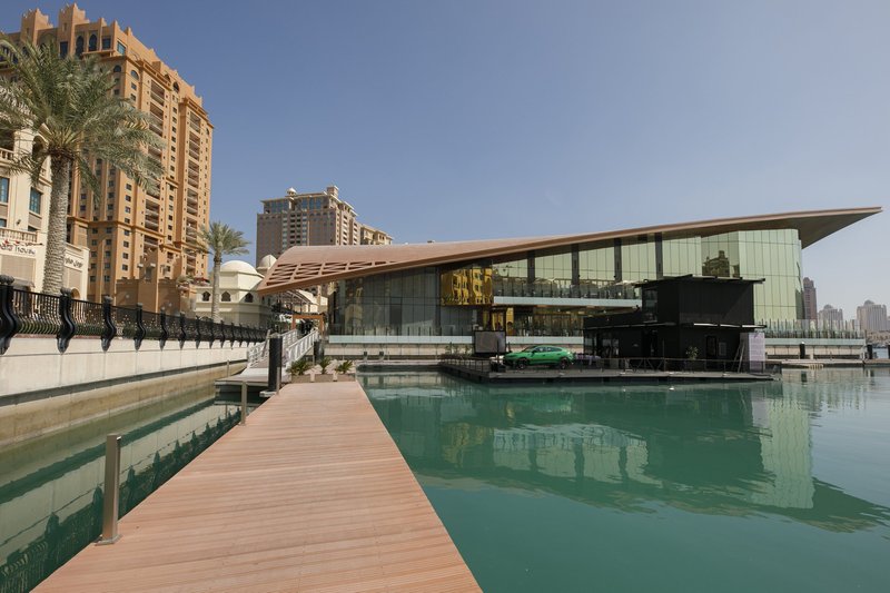 Wide shot of the Lamborghini Lounge Doha, a modern glass-walled floating structure on the waterfront with a green Urus Performante parked outside