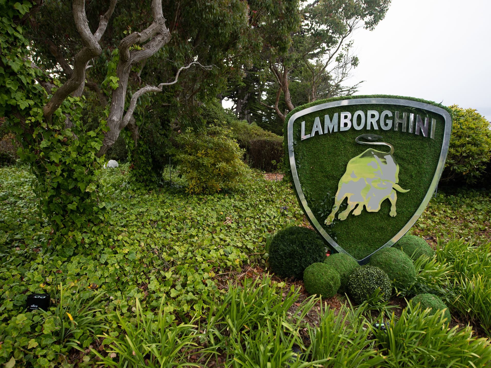 Inside Lamborghini'S Pebble Beach Playbook: How The Lounge Monterey Sells A Lifestyle, Not Just Supercars 11 Large lamborghini shield logo crafted from metal and green turf standing among lush foliage at the lounge monterey grounds