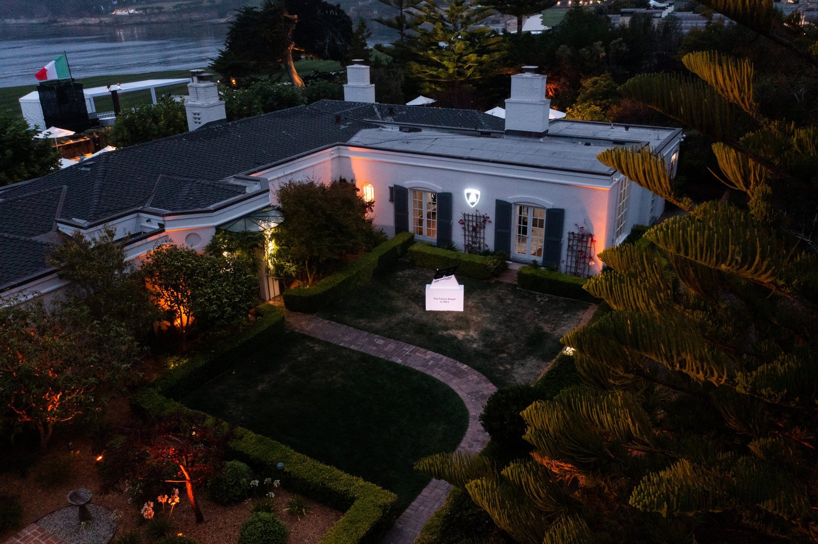 Aerial view of the Lamborghini Lounge Monterey private villa at dusk, illuminated with warm lighting and a display pavilion in the garden