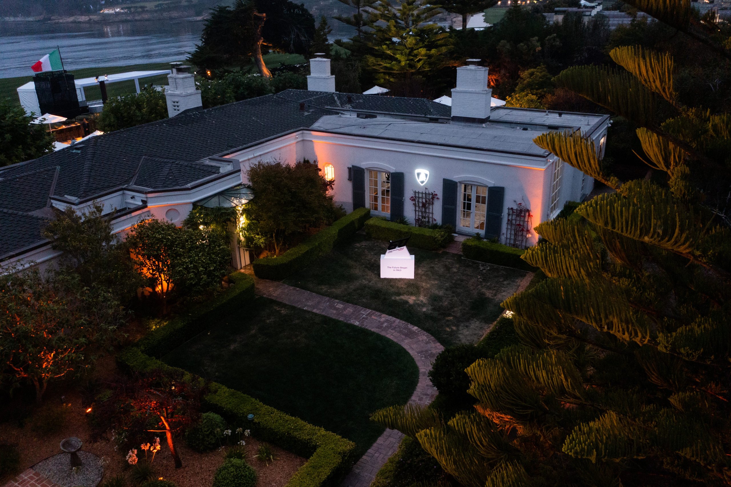 Aerial view of the lamborghini lounge monterey private villa at dusk, illuminated with warm lighting and a display pavilion in the garden