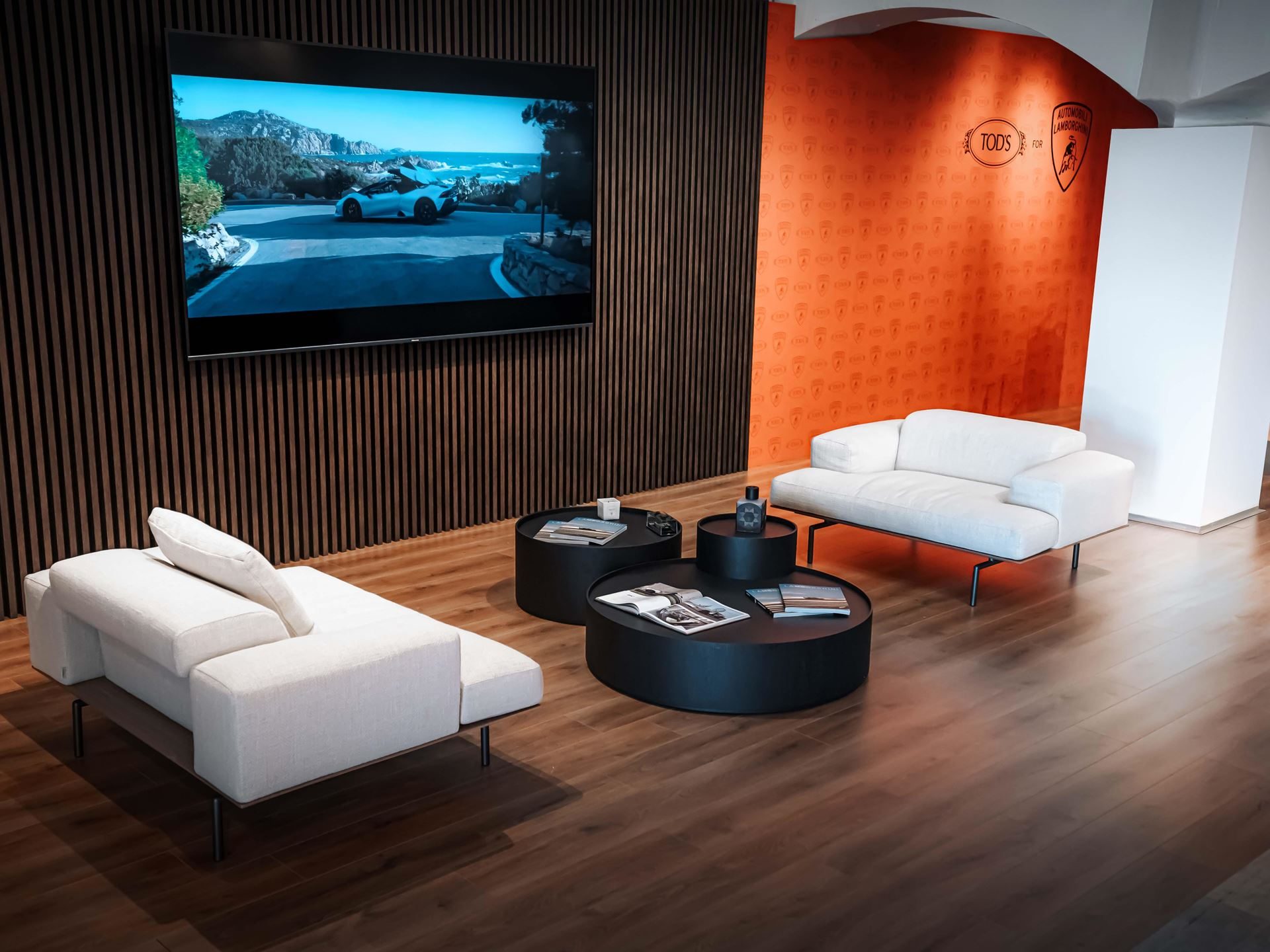 Lamborghini'S Porto Cervo Lounge Returns For A Sixth Summer As A Seaside Brand Hub 3 Modern lounge area with white sofas and large screen displaying a lamborghini revuelto, with partner logos on an orange accent wall