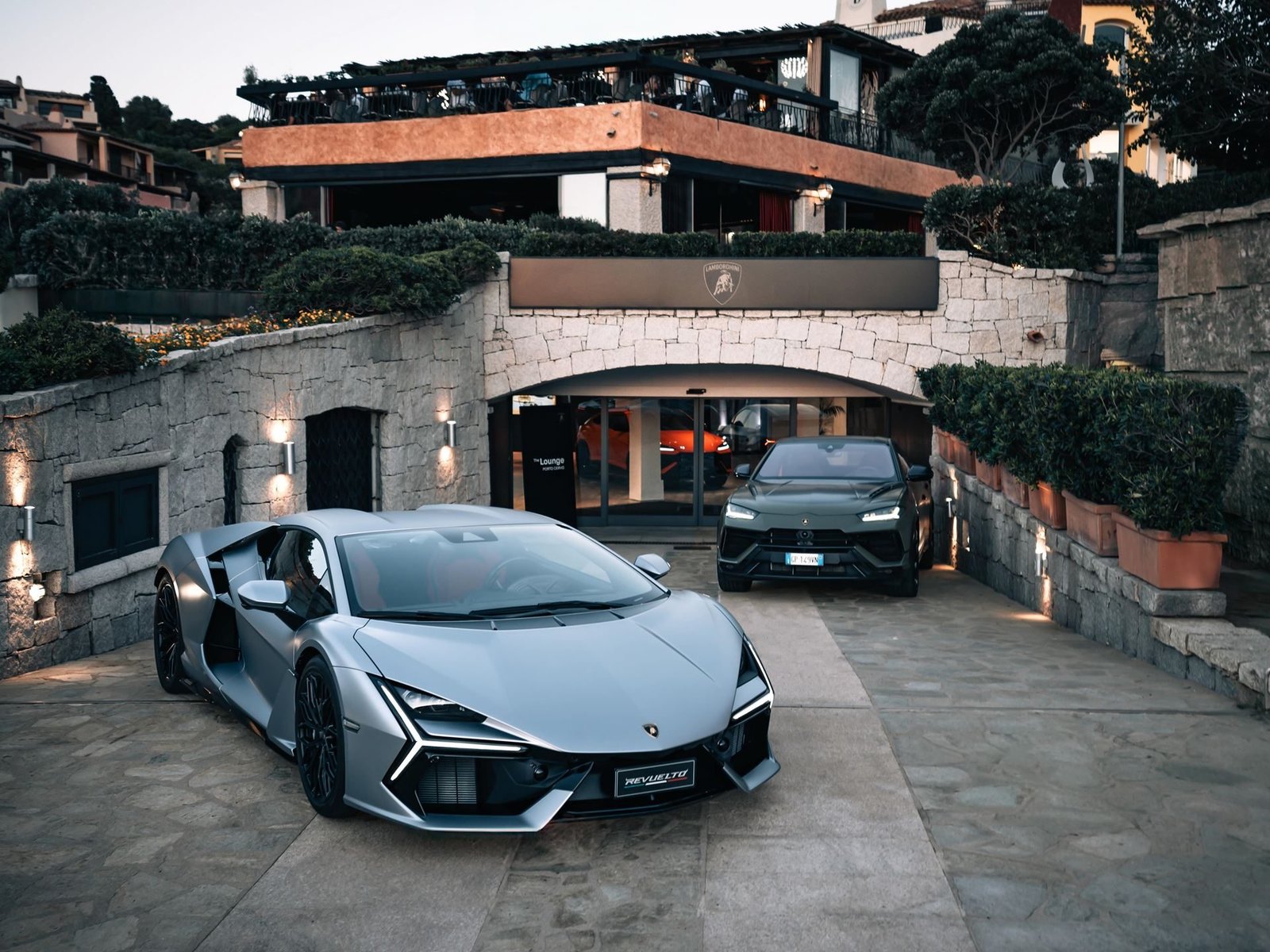 A silver Lamborghini Revuelto and dark green Lamborghini Urus parked outside The Lounge Porto Cervo at dusk
