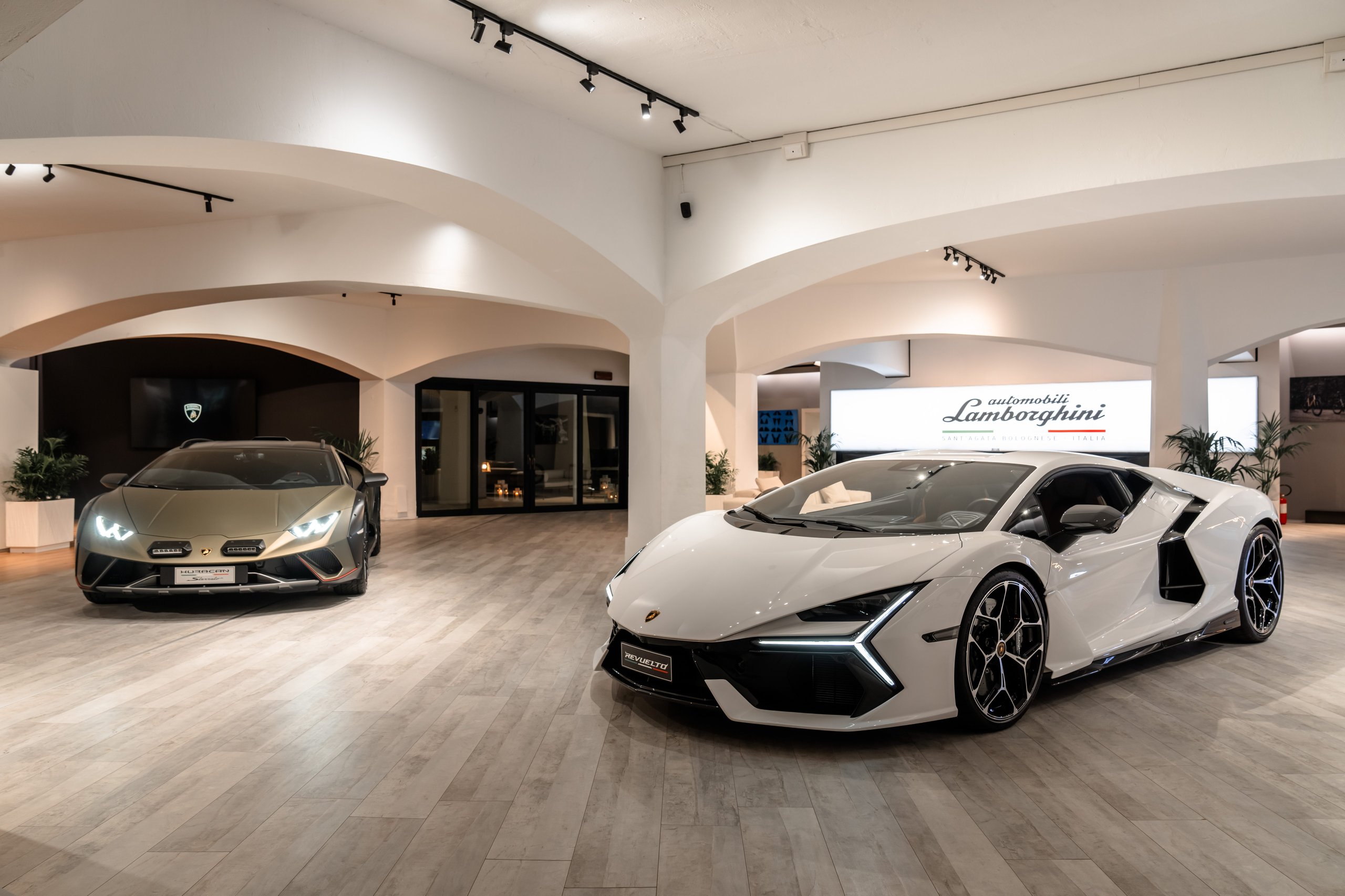 Inside Lamborghini'S Porto Cervo Lounge, Where The Revuelto Meets Its Future Owners 3 White lamborghini revuelto and gold huracán displayed in a brightly lit showroom with a large lamborghini logo