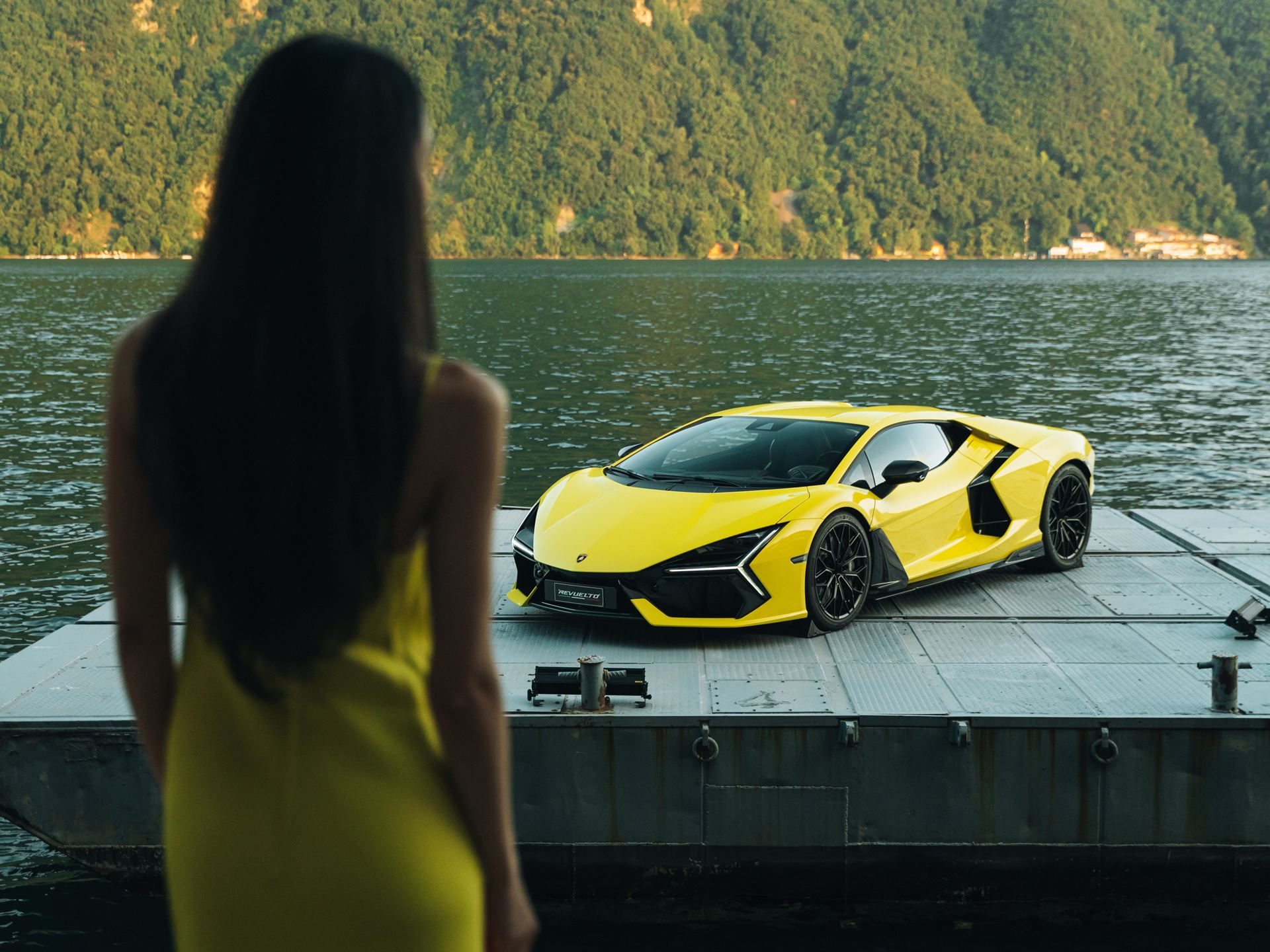 Lamborghini Lugano Turned Its First Birthday Into A Lakeside Stage For The Urus Se'S Swiss Debut 3 A woman in a yellow dress looks out at a yellow lamborghini revuelto displayed on a floating platform on lake lugano with mountains in the background