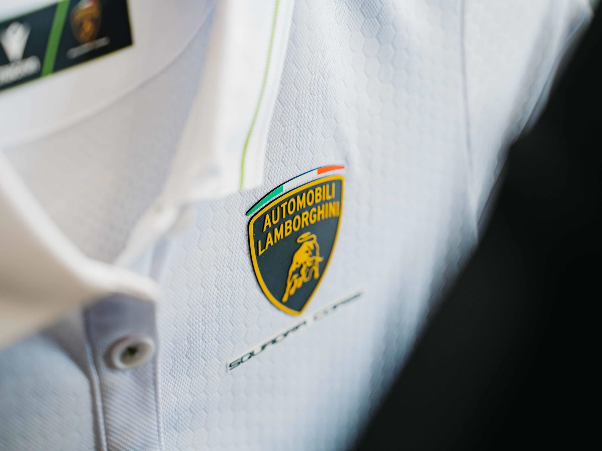 Lamborghini Taps Macron To Outfit Squadra Corse For Three Seasons, Replica Collection Included 9 Close-up of a white polo shirt featuring the automobili lamborghini squadra corse shield logo with visible fabric texture