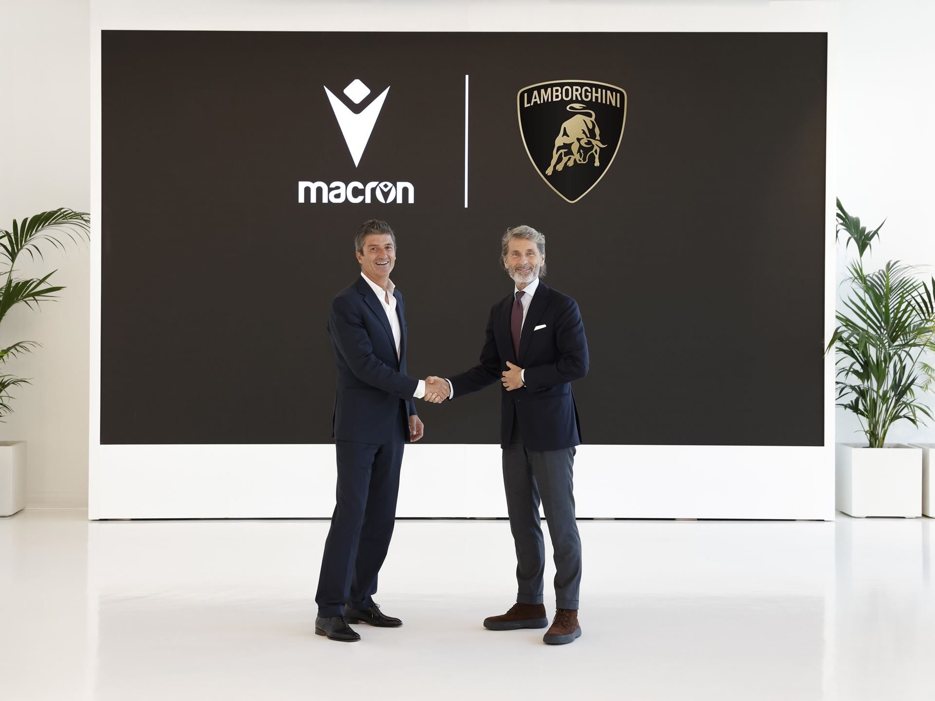 Lamborghini Taps Macron To Outfit Squadra Corse For Three Seasons, Replica Collection Included 3 Two executives shake hands in front of a screen displaying macron and lamborghini logos, formalizing the partnership