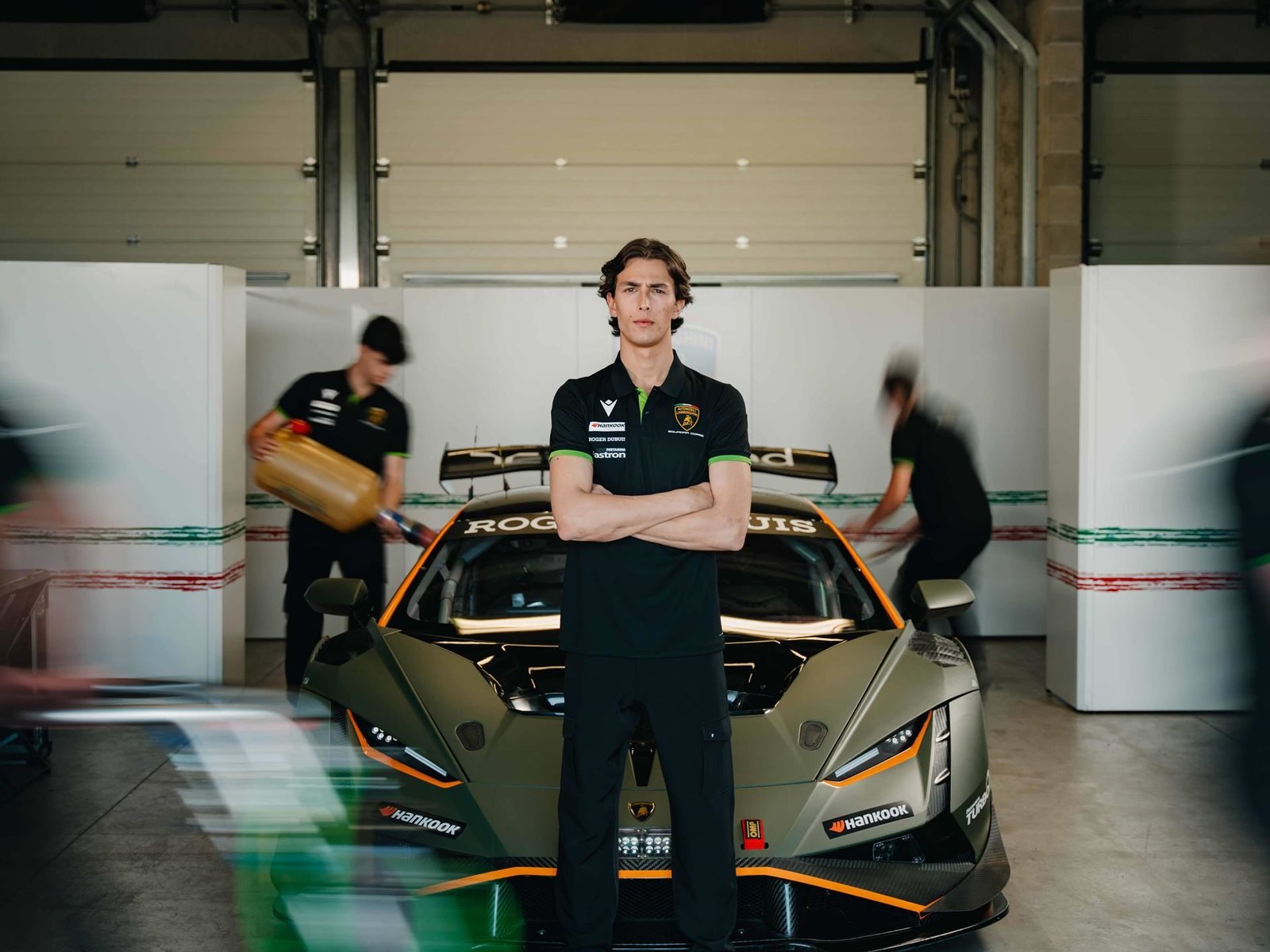 A Lamborghini Squadra Corse driver stands with arms crossed in front of a Huracán GT3 EVO2 in a pit garage, wearing Macron team apparel