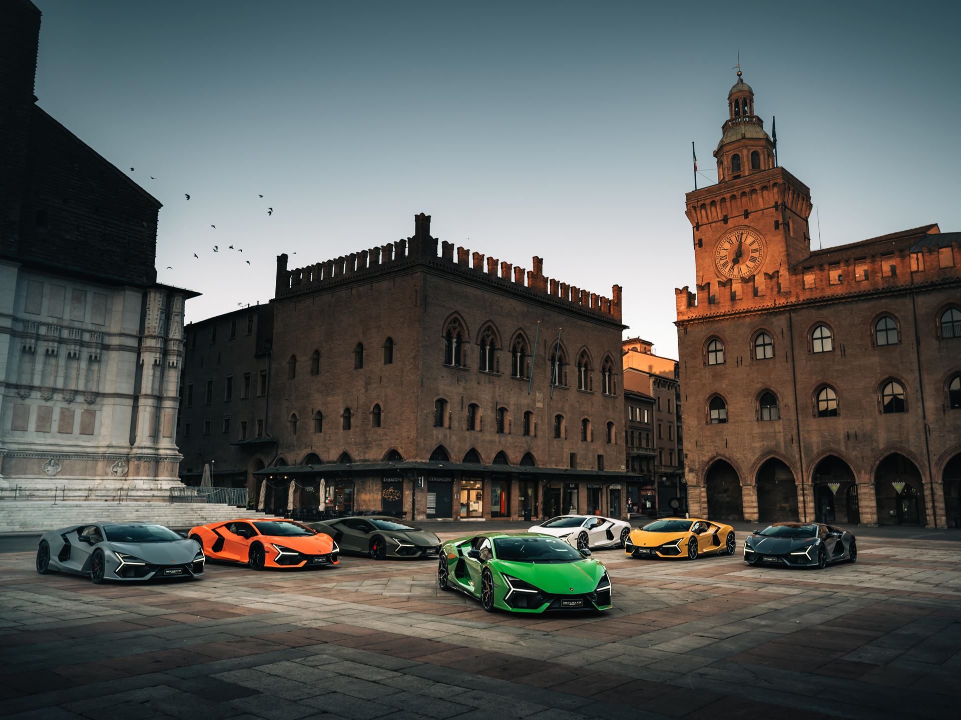 Why Lamborghini'S 'Made In Italy' Day Matters More Than National Pride 9 Collection of lamborghini supercars and suvs including a green revuelto parked in a historic italian city square with ancient buildings and clock tower