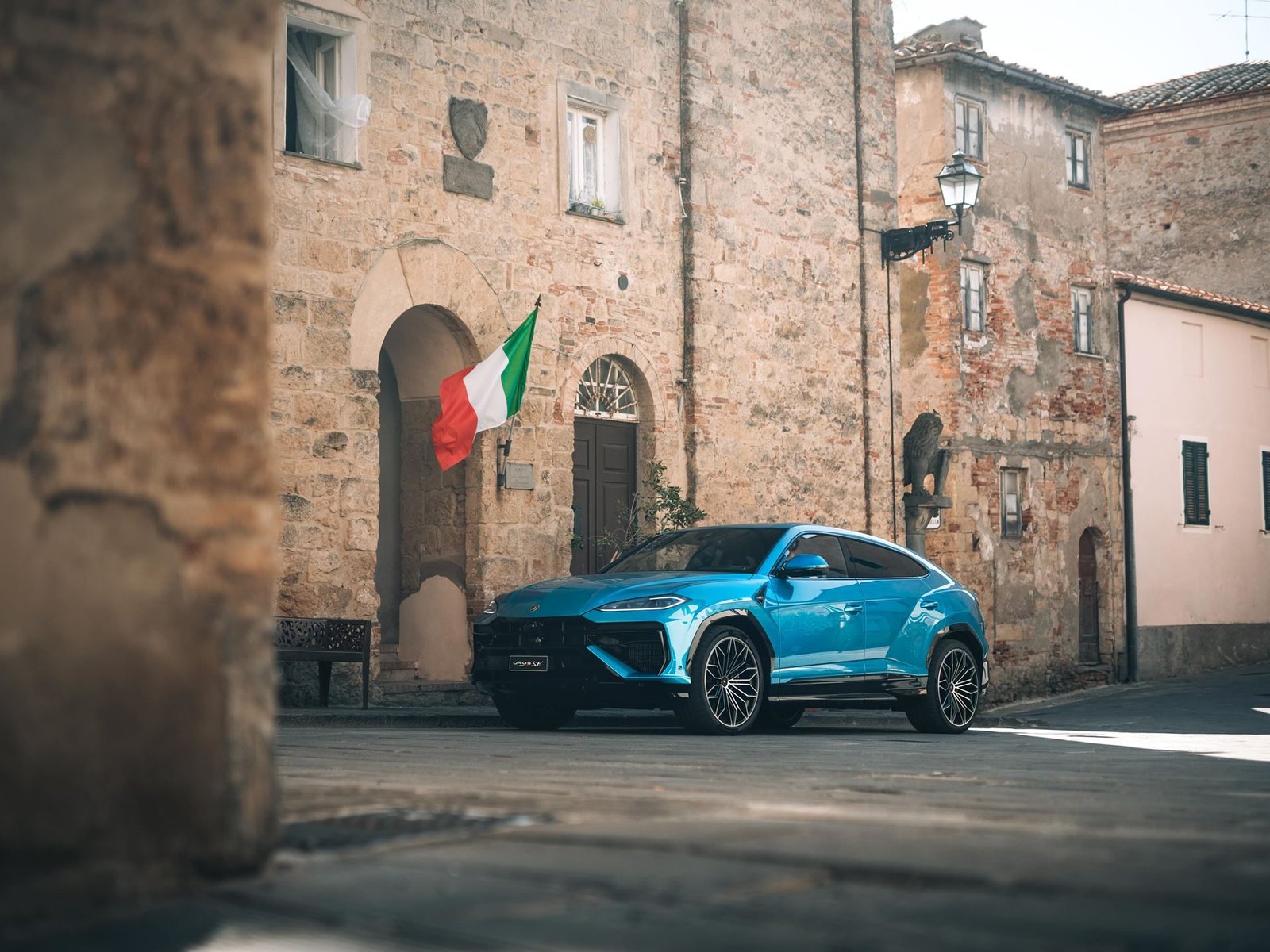 Blue Lamborghini Urus parked on a cobblestone street in a historic Italian town with an Italian flag hanging from a building