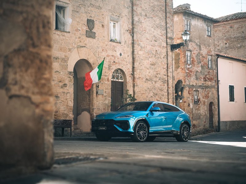 Blue Lamborghini Urus parked on a cobblestone street in a historic Italian town with an Italian flag hanging from a building