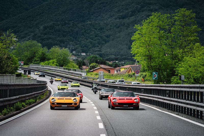 A convoy of classic Lamborghini Miuras in orange and red driving along a winding mountain road through lush green Italian hills