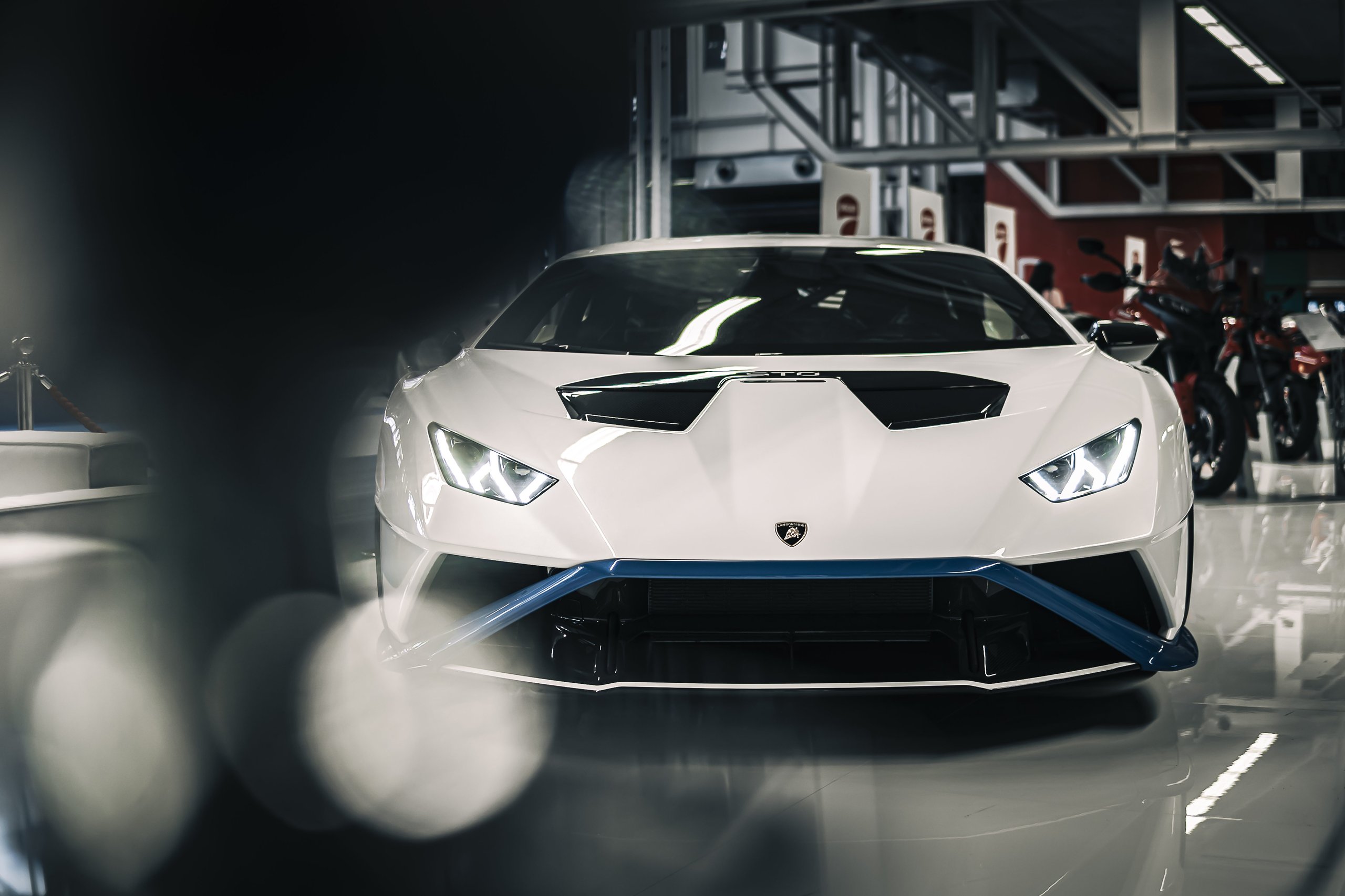 Four Models, Three Executives, One Message: Lamborghini Used Motor Valley Fest 2022 To Map Its Entire Future 7 Front view of a white lamborghini huracán sto with blue accents and headlights on at an indoor exhibition