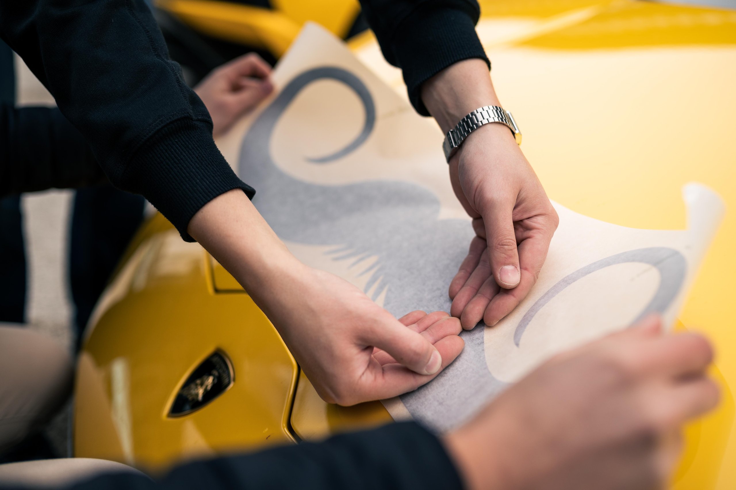 Over 800 Lamborghinis Wear Mustaches For Movember, And Bologna Fc Joins The Parade 7 Two pairs of hands carefully positioning a large mustache decal onto the yellow hood of a lamborghini