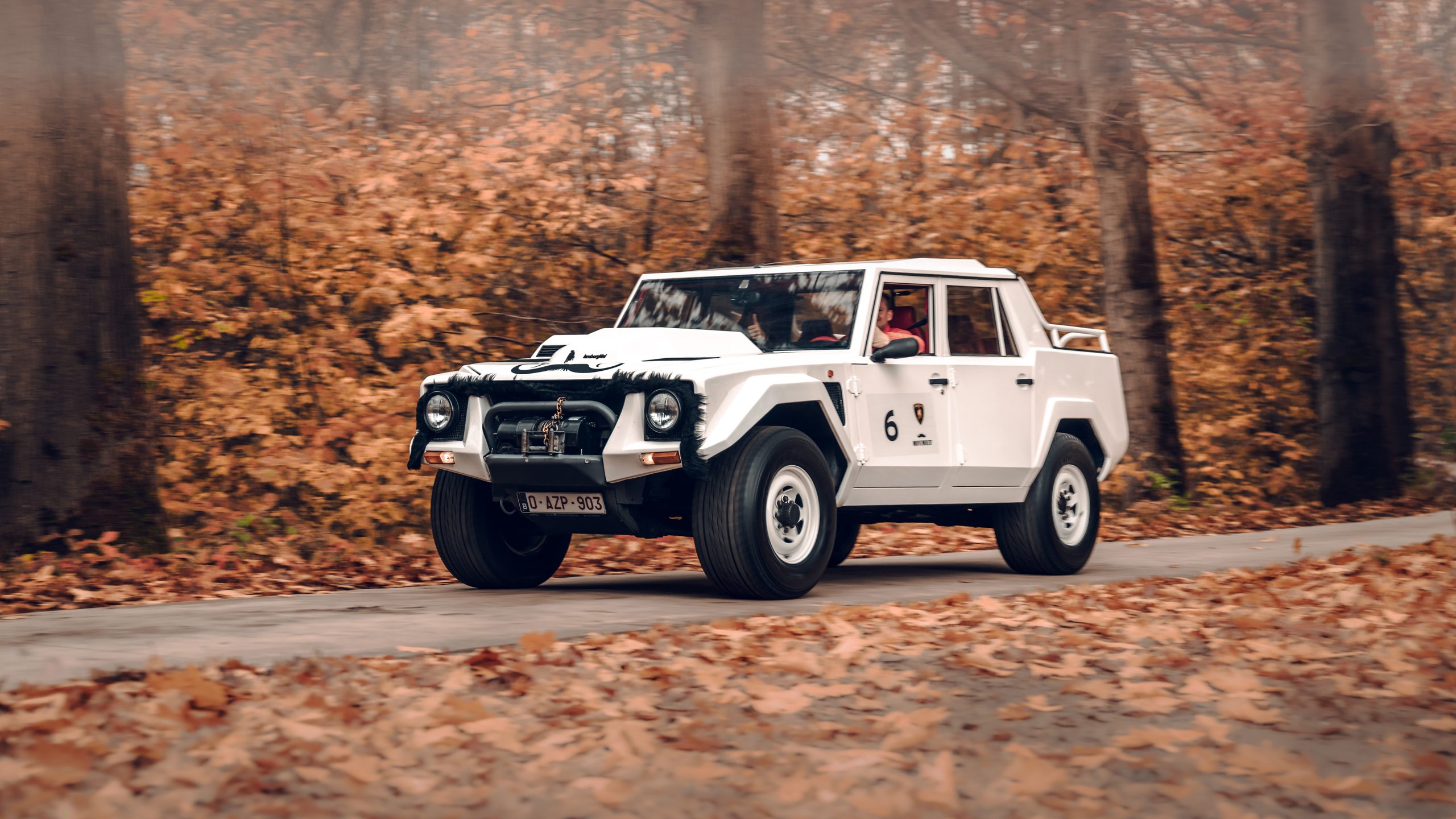 A white lamborghini lm002 driving on a winding road through an autumn forest with fallen leaves