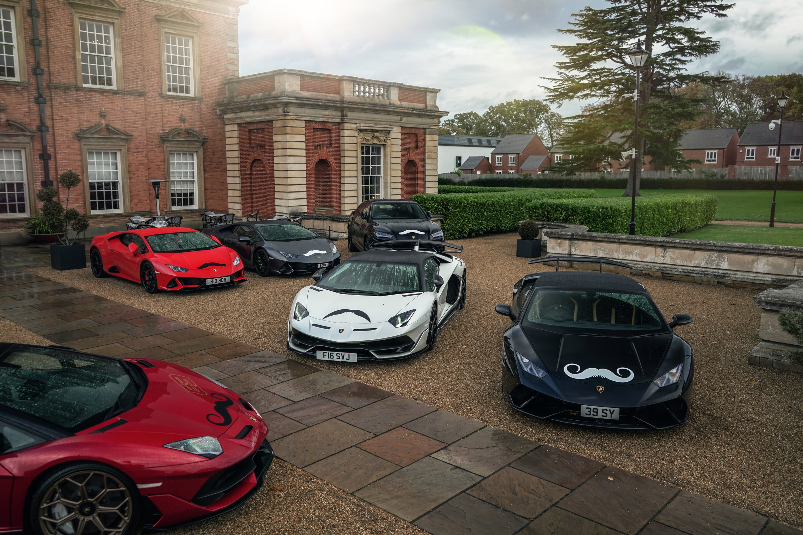 A collection of lamborghini huracáns, aventadors, and urus models with movember mustache decals parked in front of a grand estate