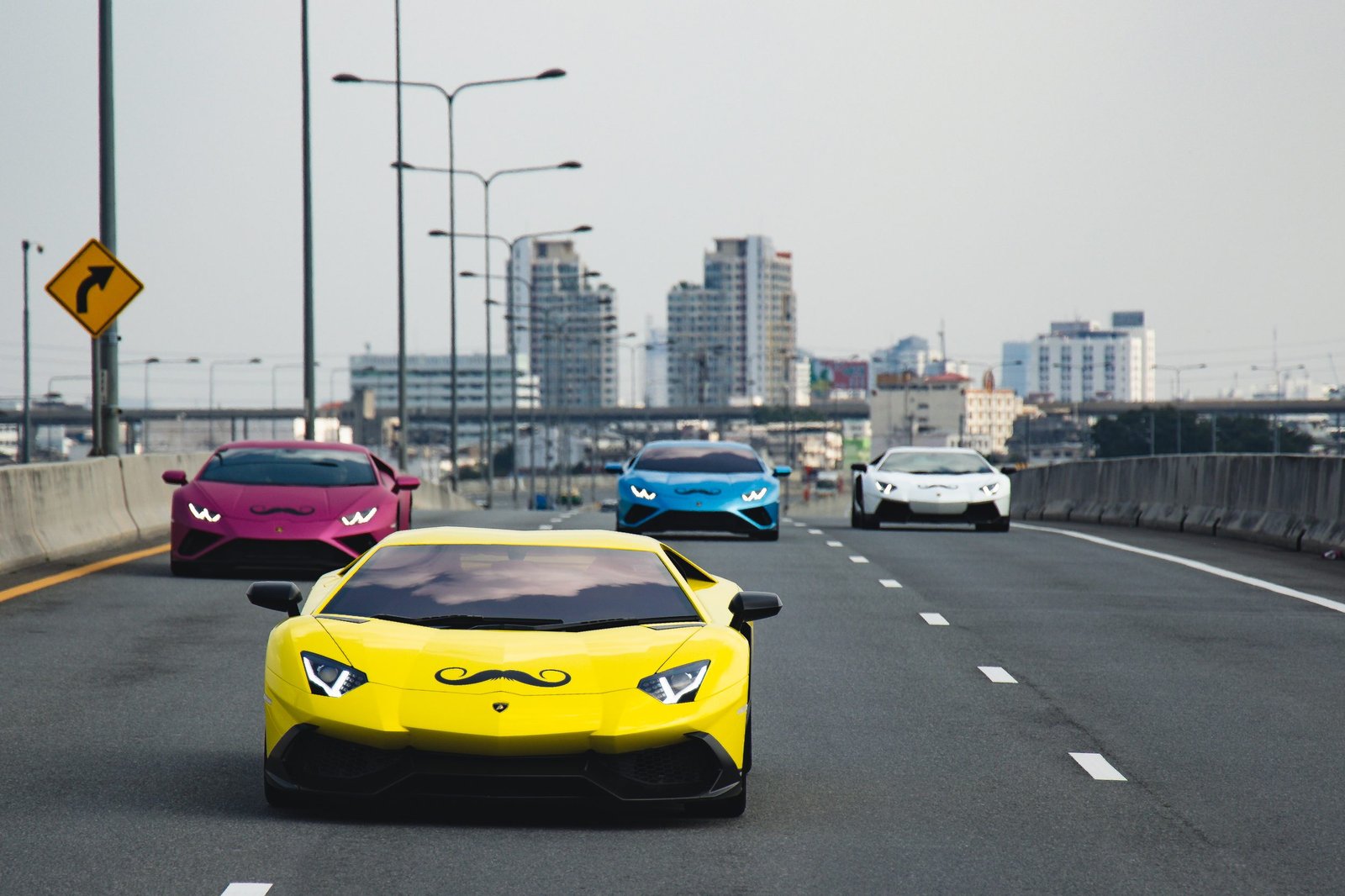 A yellow Lamborghini supercar with a Movember mustache decal leads a colorful convoy of Lamborghinis on a highway with a city skyline in the background