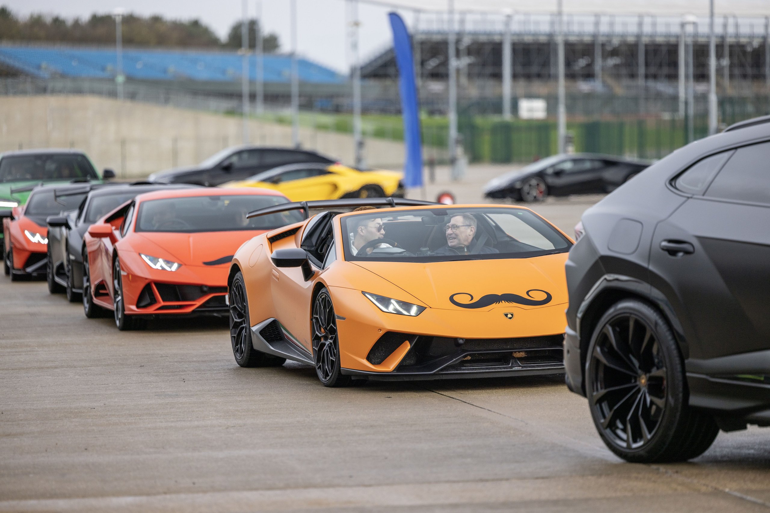 A line of lamborghini cars including an orange huracán with movember mustache decal and a grey urus driving on the silverstone track