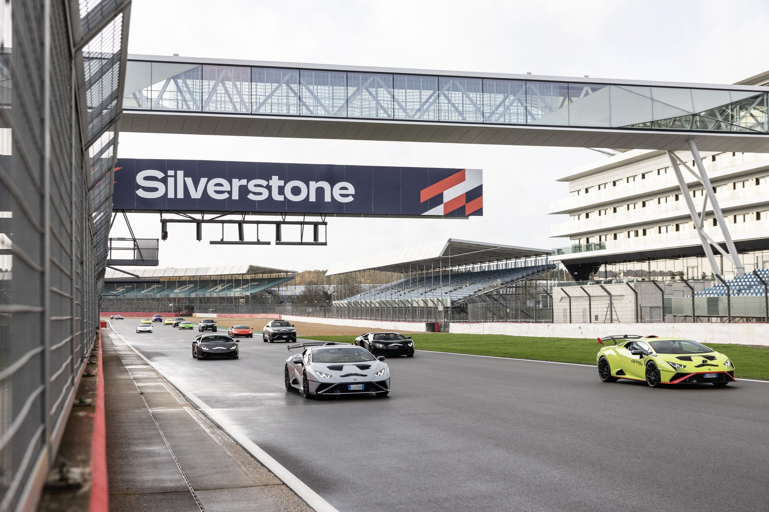 Line of lamborghini huracáns and urus suvs driving on the silverstone racetrack with grandstands visible in background
