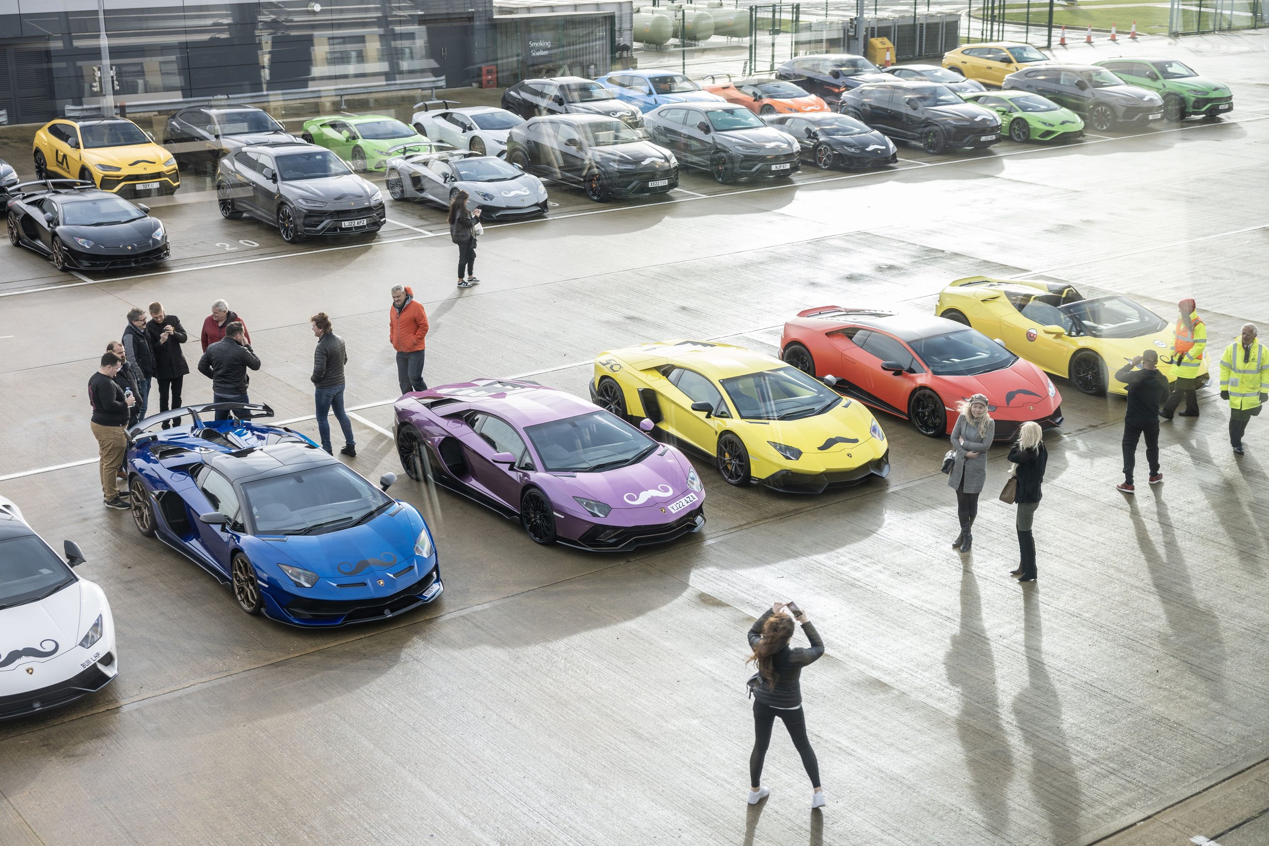 Overhead shot of lamborghini huracáns, aventadors, and urus suvs with movember mustache decals parked in the silverstone paddock with owners mingling