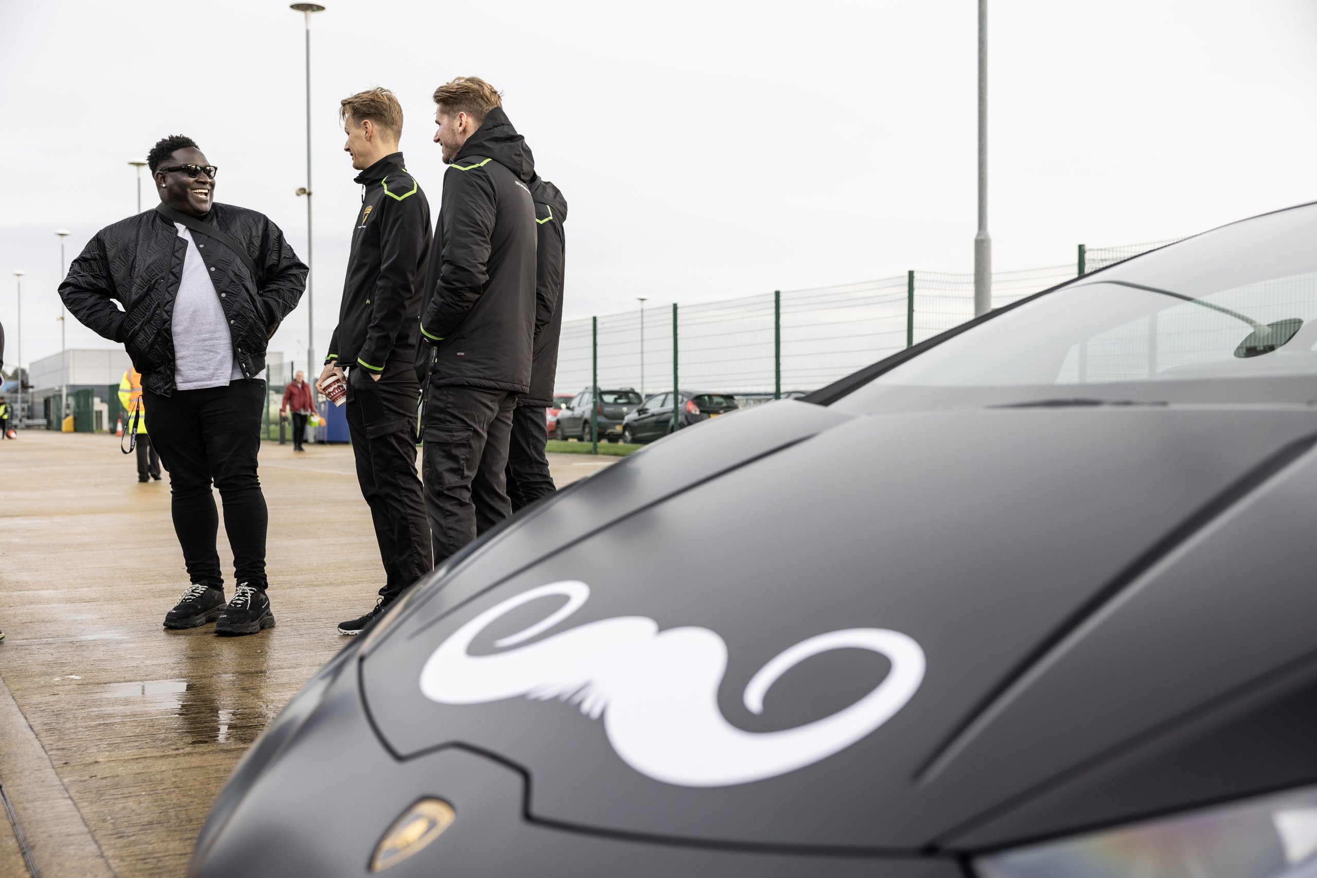 Close-up of a black lamborghini urus hood with white movember mustache decal and group of participants in background