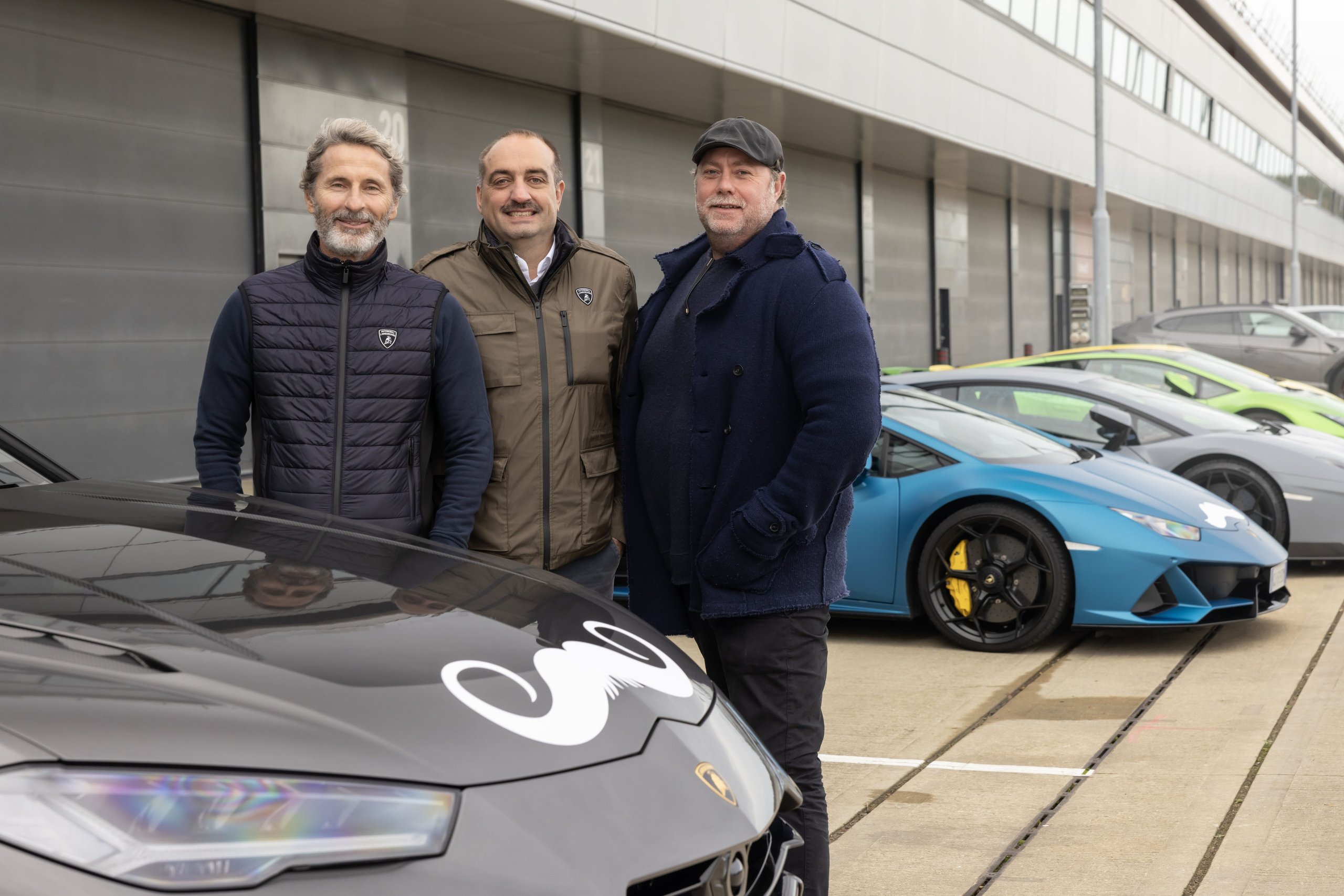 Three men smiling in front of a grey lamborghini urus with movember mustache decal and a blue huracán at the silverstone event