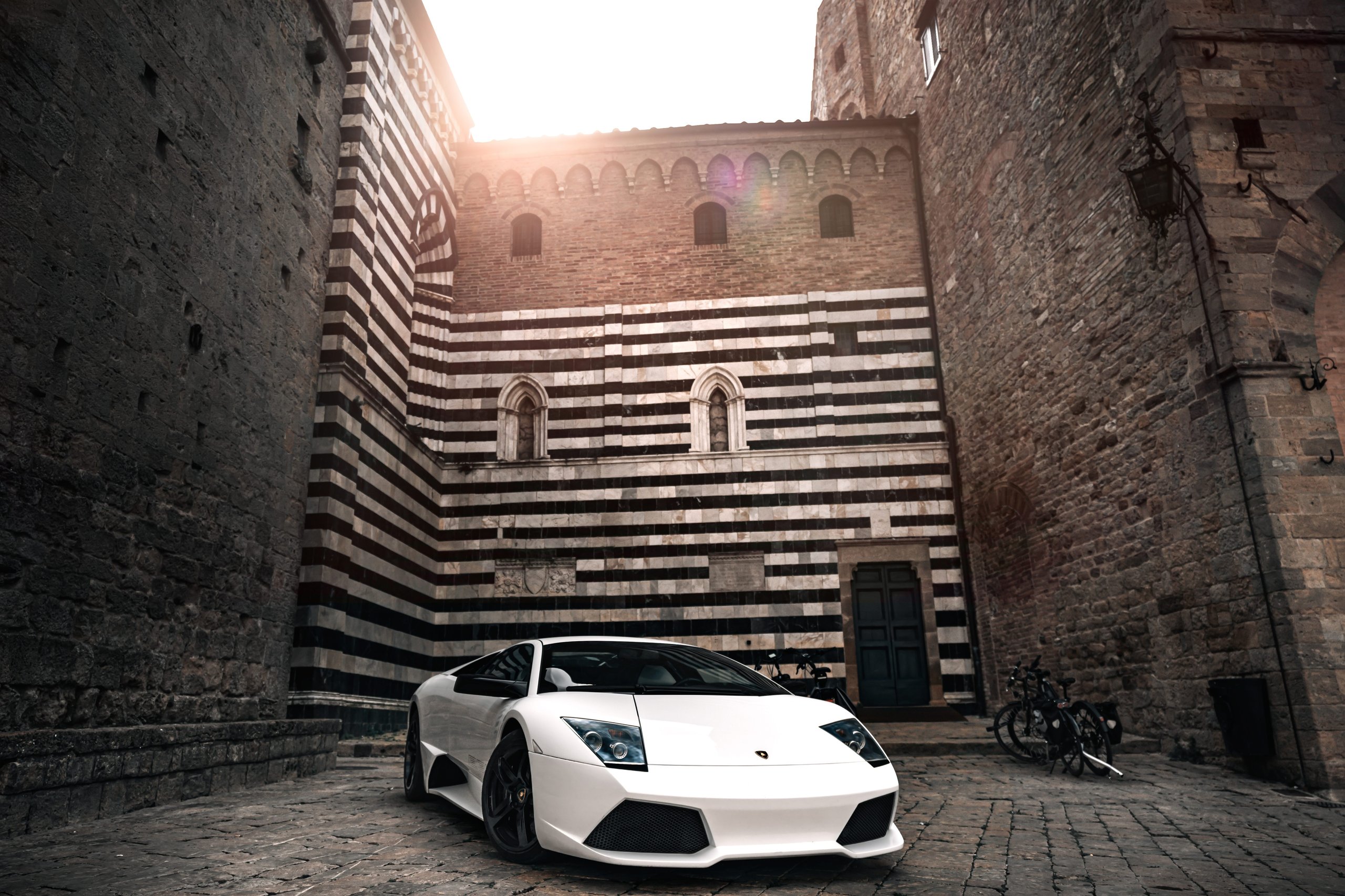 The Murciélago Was Lamborghini'S Last V12 You Could Row Yourself, And That Changes Everything About How We Remember It 3 White lamborghini murciélago parked on a cobblestone street in front of a historic building with striped masonry
