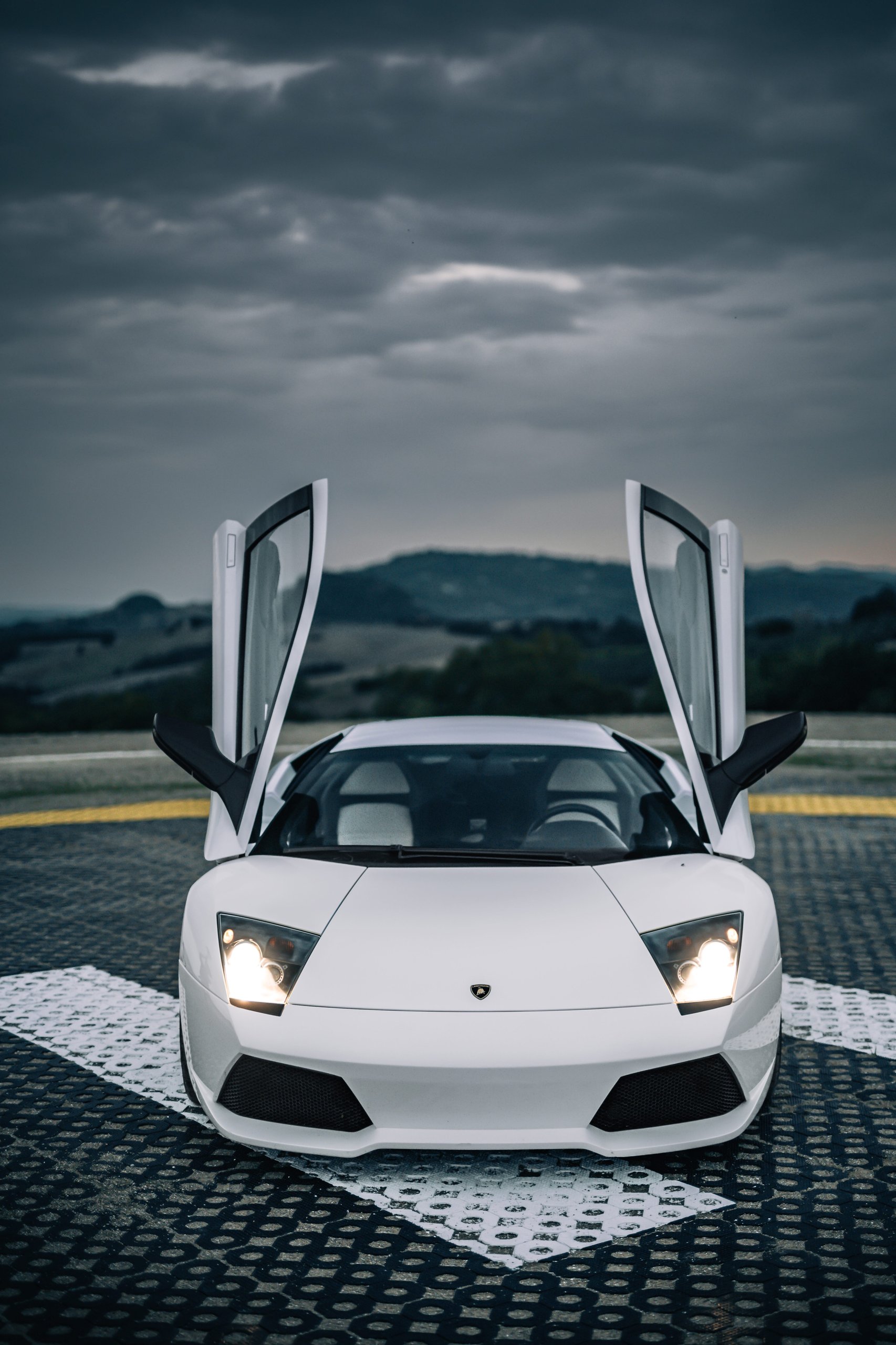 The Murciélago Was Lamborghini'S Last V12 You Could Row Yourself, And That Changes Everything About How We Remember It 9 White lamborghini murciélago with scissor doors open, parked on a helipad under a cloudy sky