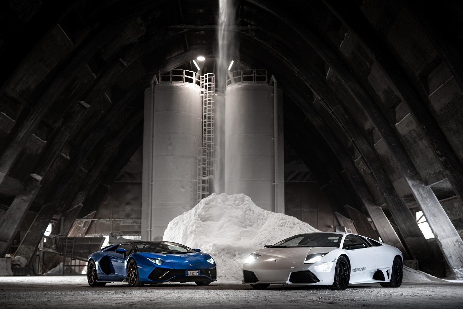 White Lamborghini Murciélago and blue Lamborghini Aventador parked side by side in an industrial setting, illustrating the V12 lineage