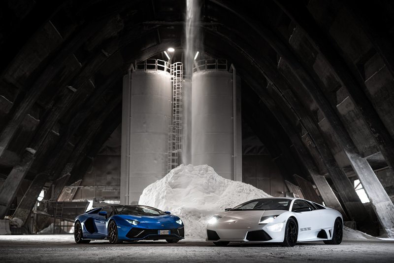 White Lamborghini Murciélago and blue Lamborghini Aventador parked side by side in an industrial setting, illustrating the V12 lineage