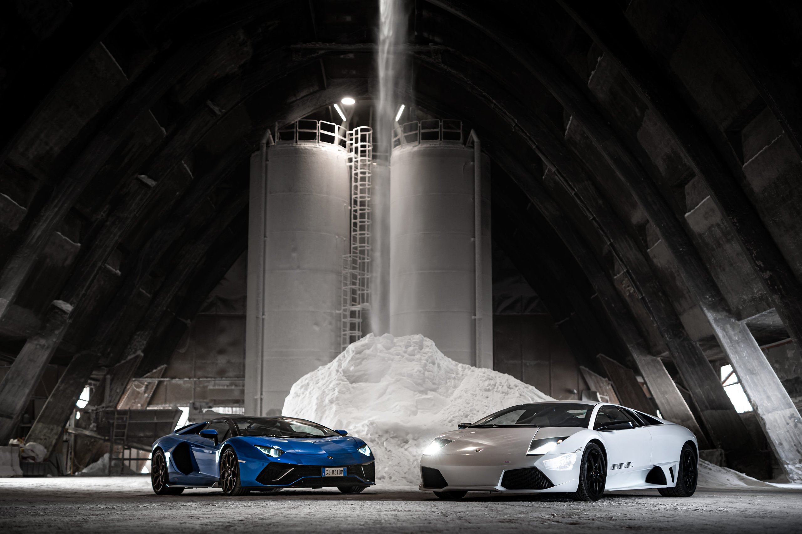 White lamborghini murciélago and blue lamborghini aventador parked side by side in an industrial setting, illustrating the v12 lineage