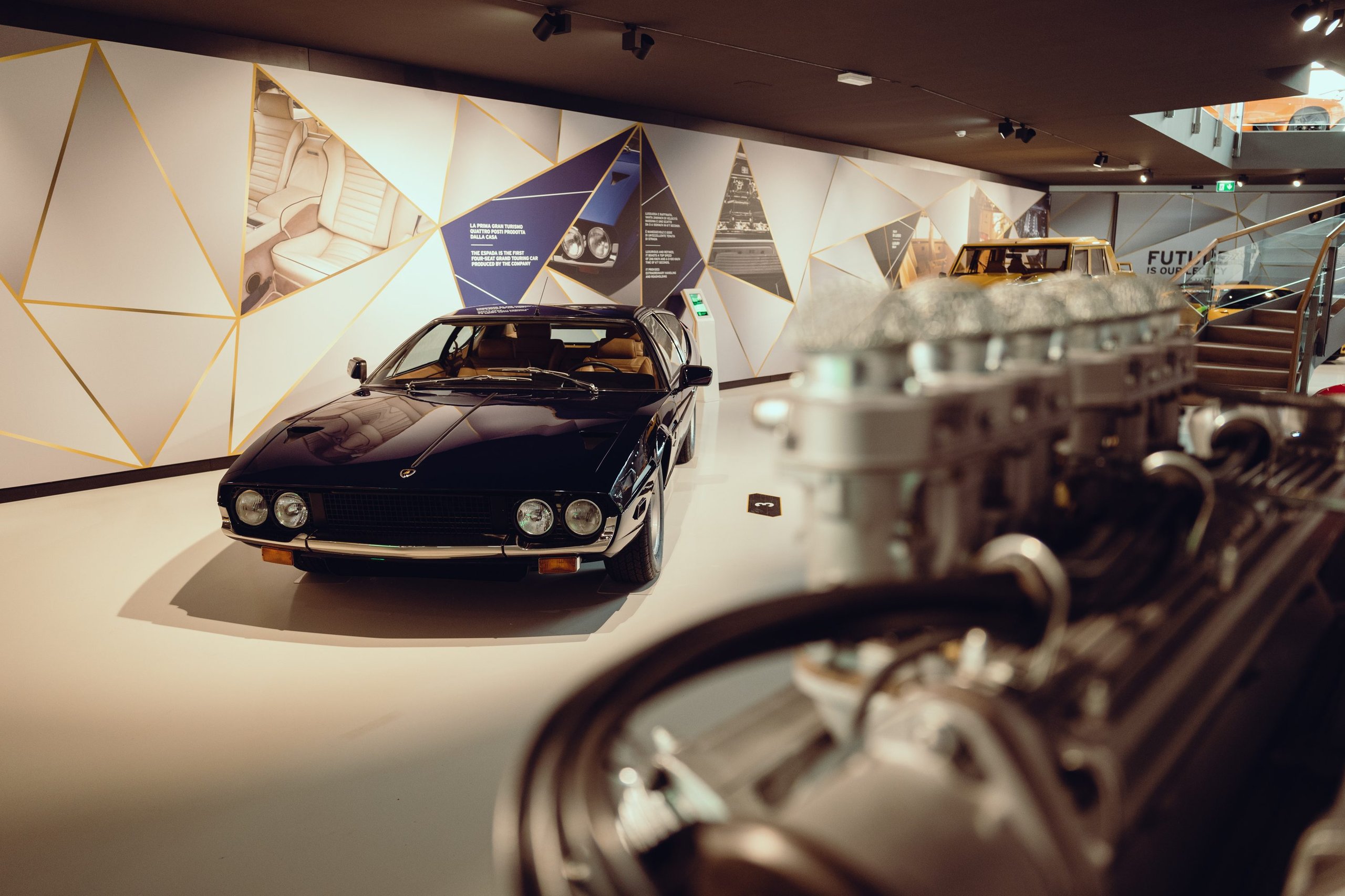 Dark blue lamborghini jarama displayed in the museum with a v12 engine visible in the foreground