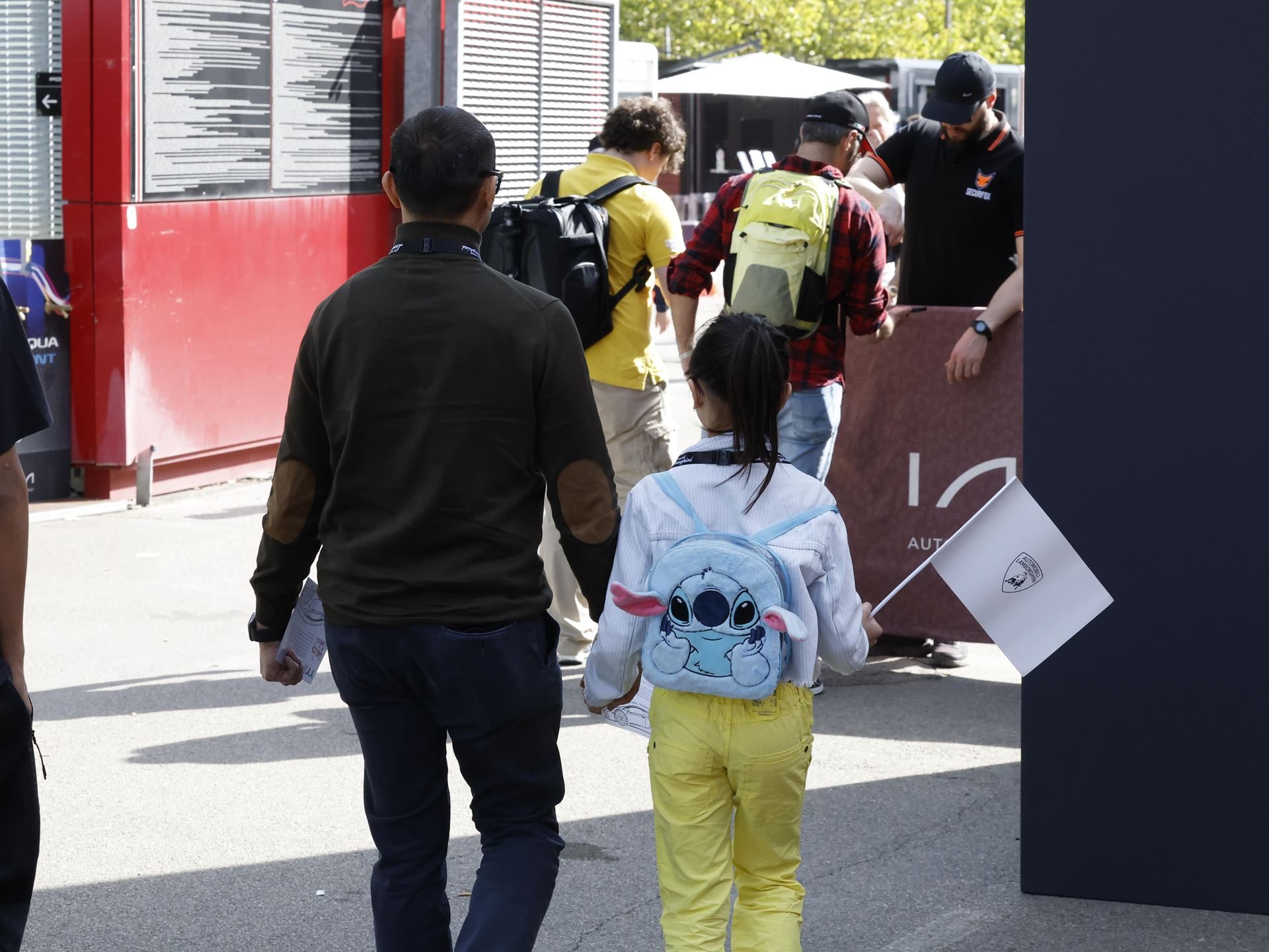 736 Parents In Sant'Agata: Inside Lamborghini'S Workplace Support Package For Employee Families 3 A father and daughter walking into a lamborghini event, the daughter carrying a small white flag with the lamborghini logo