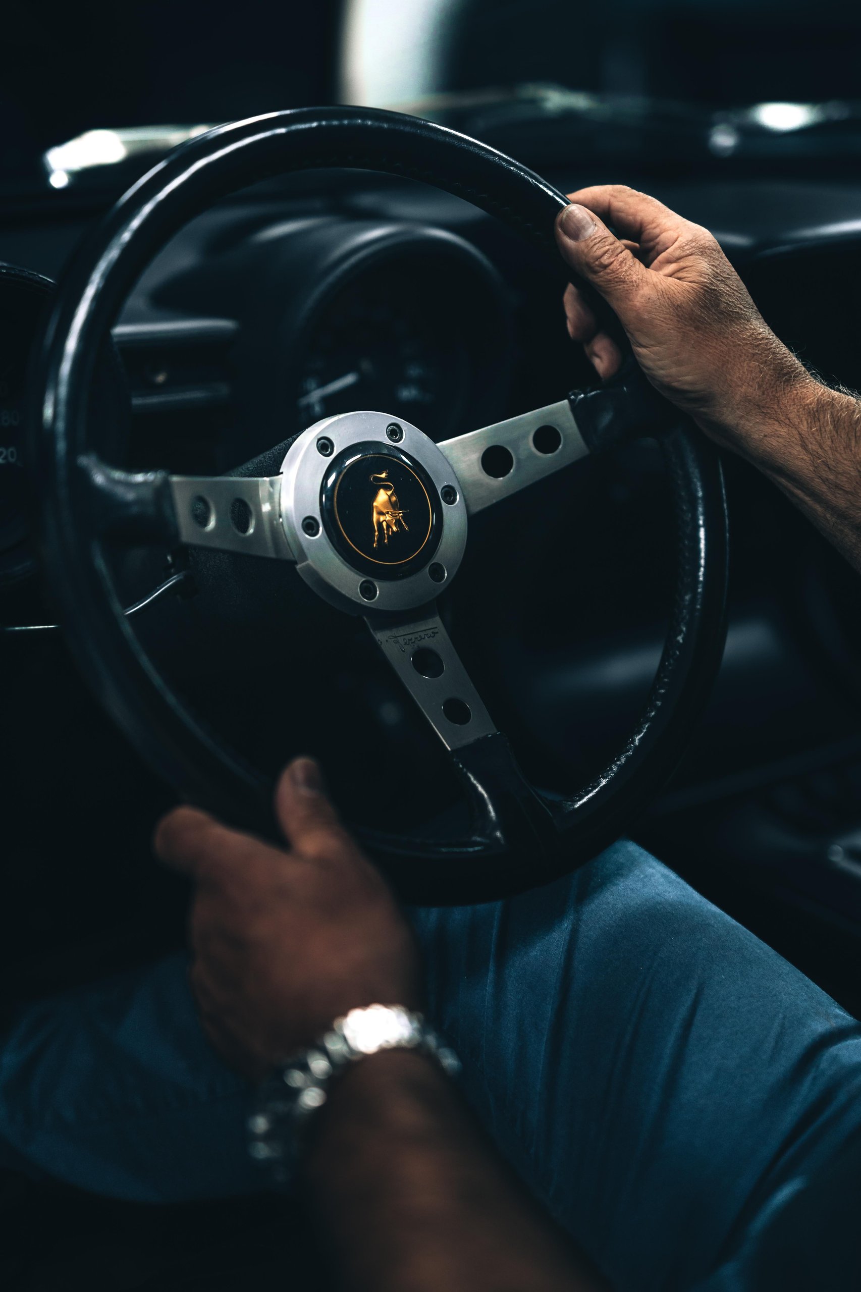 Close-up of hands gripping the classic three-spoke steering wheel of a Lamborghini Miura with the iconic bull logo on the horn button