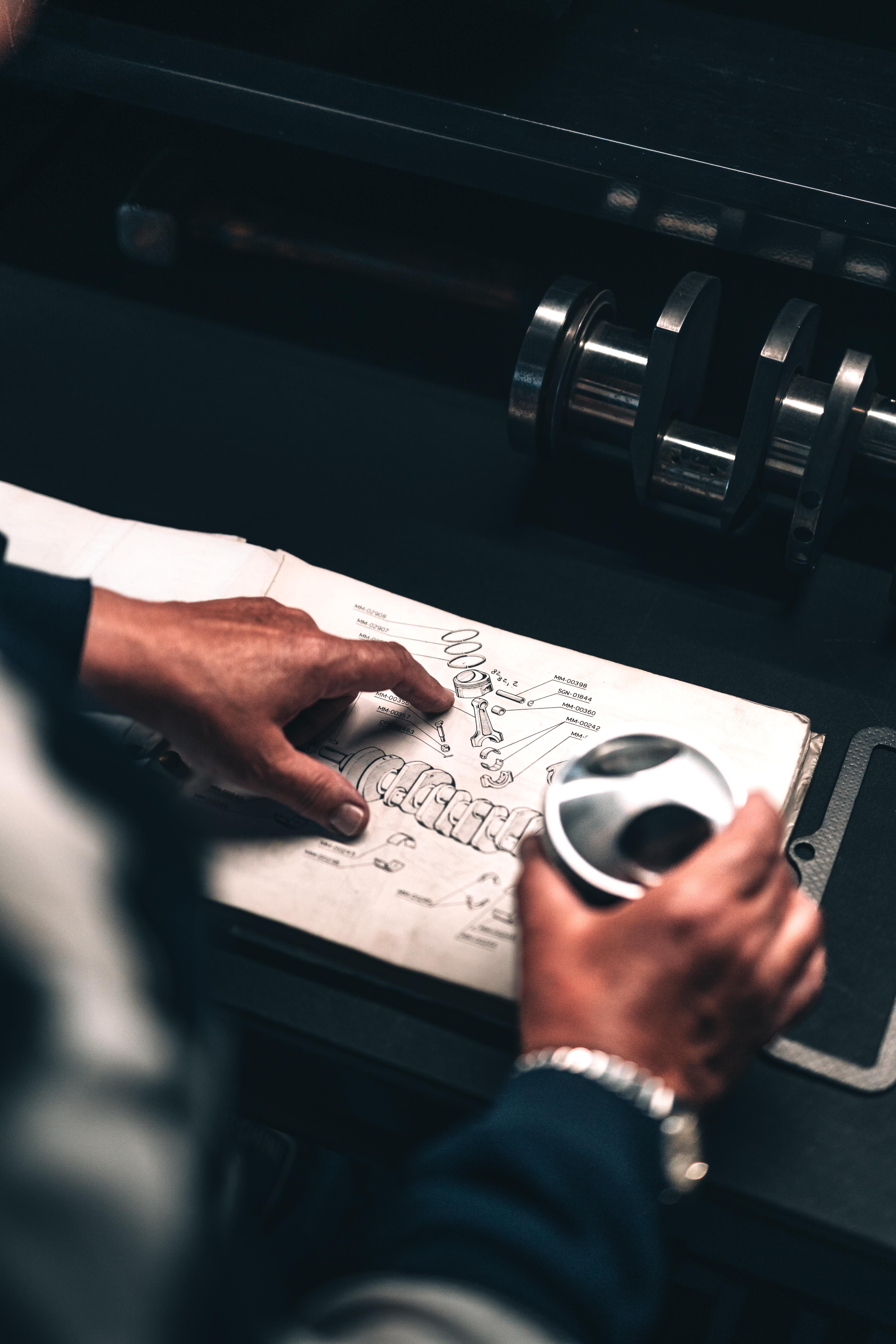 Overhead view of hands pointing at a technical drawing of engine components with a metal part and crankshaft visible