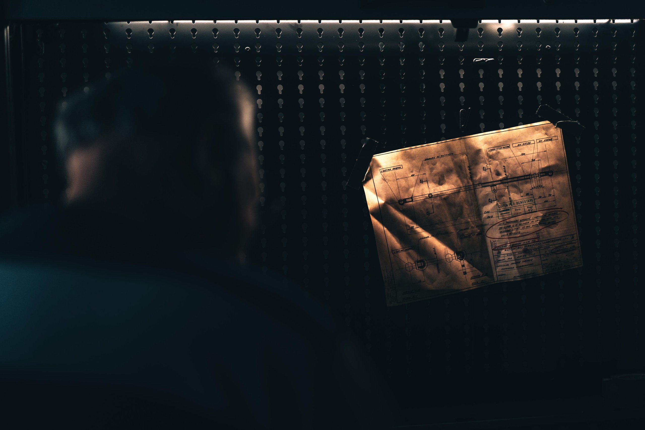Blurred figure examining an illuminated aged blueprint hanging on a tool board in the Polo Storico workshop