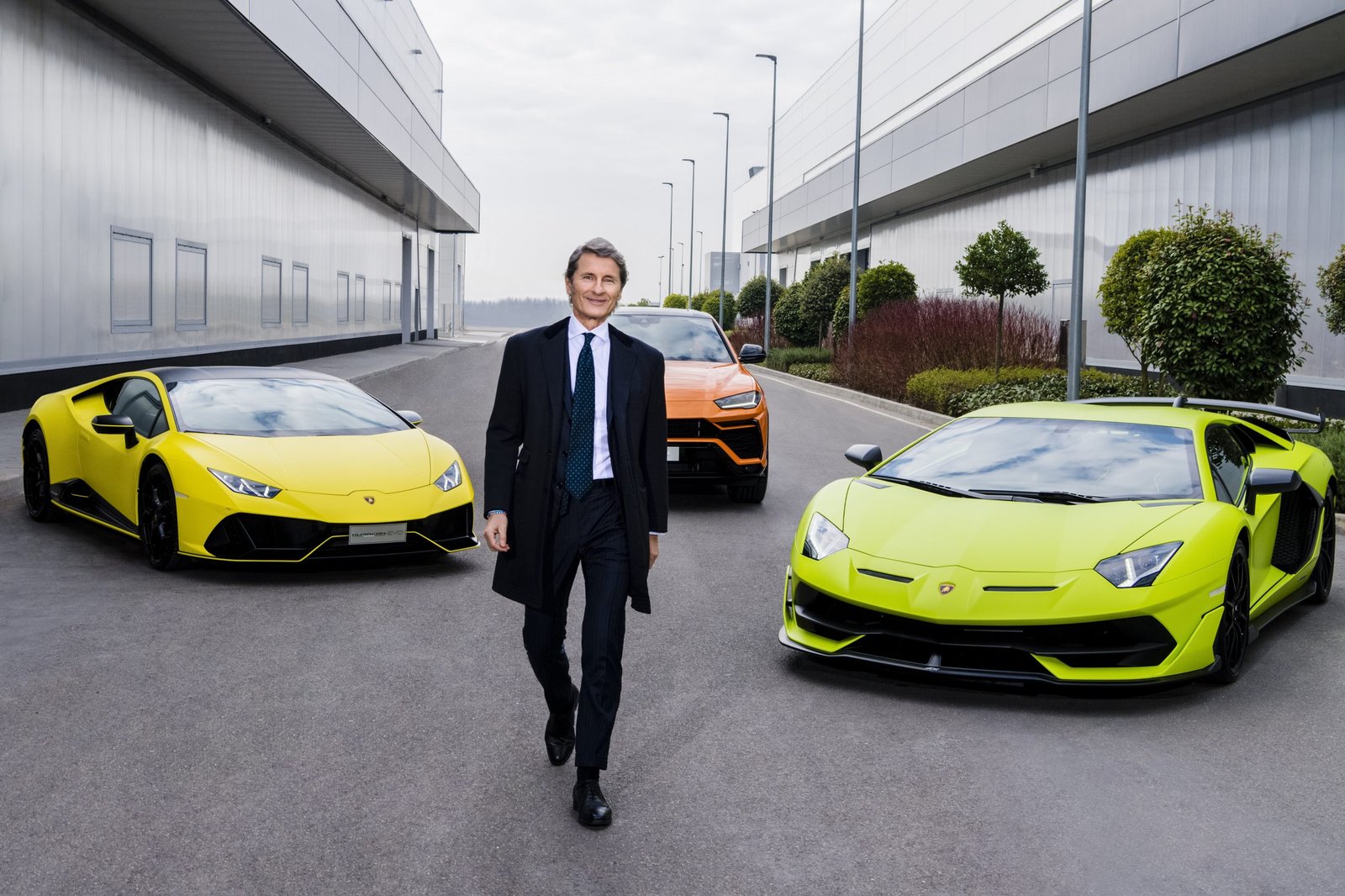 Stephan Winkelmann walking alongside a yellow Lamborghini Huracán, green Aventador, and orange Urus outside a modern Lamborghini building