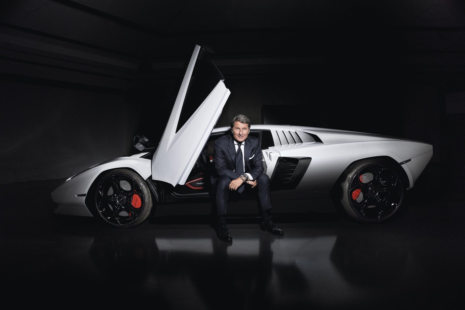 Stephan Winkelmann, CEO of Lamborghini, seated on the sill of a white Lamborghini Countach LPI 800-4 with its scissor door open