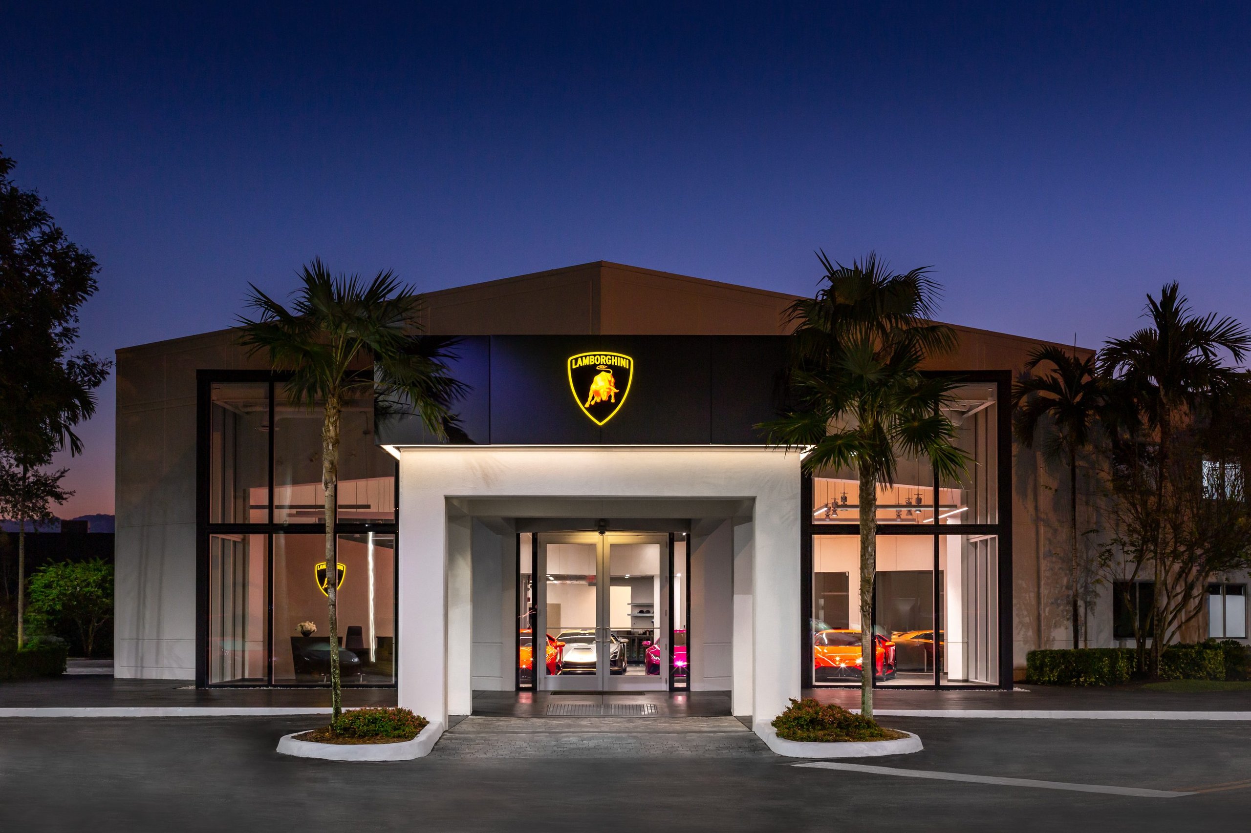 California And Florida Drive Lamborghini'S Record 2022 U.s. Sales As Two Showrooms Debut A New Retail Identity 3 Exterior of a modern lamborghini showroom at dusk with illuminated shield logo, large glass windows, and palm trees flanking the entrance