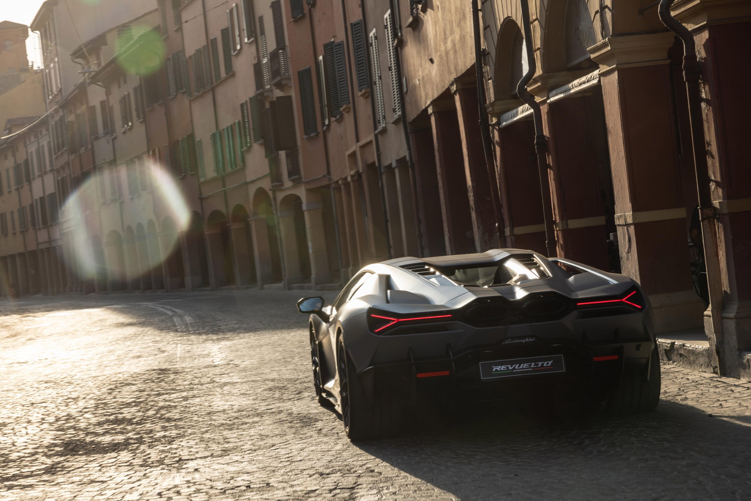Lamborghini Sent The Revuelto Across Italy To Prove A 1,001 Hp Hybrid Can Still Be A Proper V12 Flagship 3 Dark grey lamborghini revuelto driving down a historic cobblestone street with sunlight flaring behind