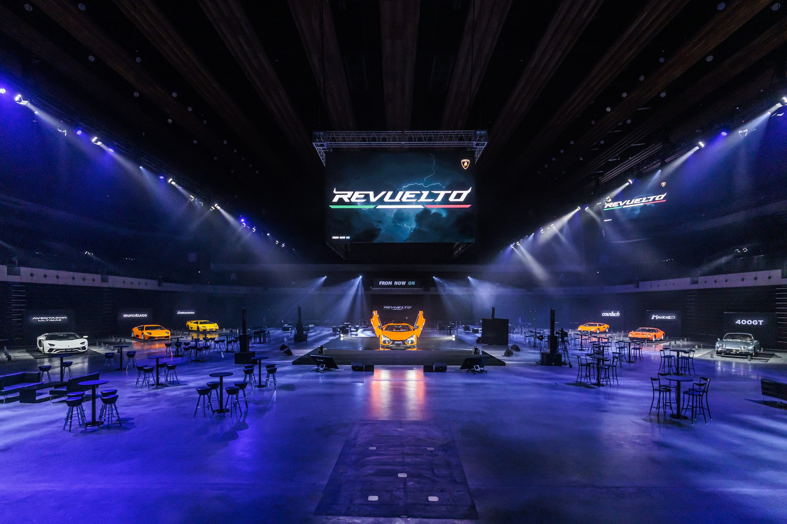 Lamborghini Brought Six Decades Of V12 History To Tokyo'S Ariake Arena, Then Unveiled What Comes Next 3 Wide shot of the lamborghini revuelto japan premiere event showing multiple heritage v12 models under dramatic blue and purple lighting
