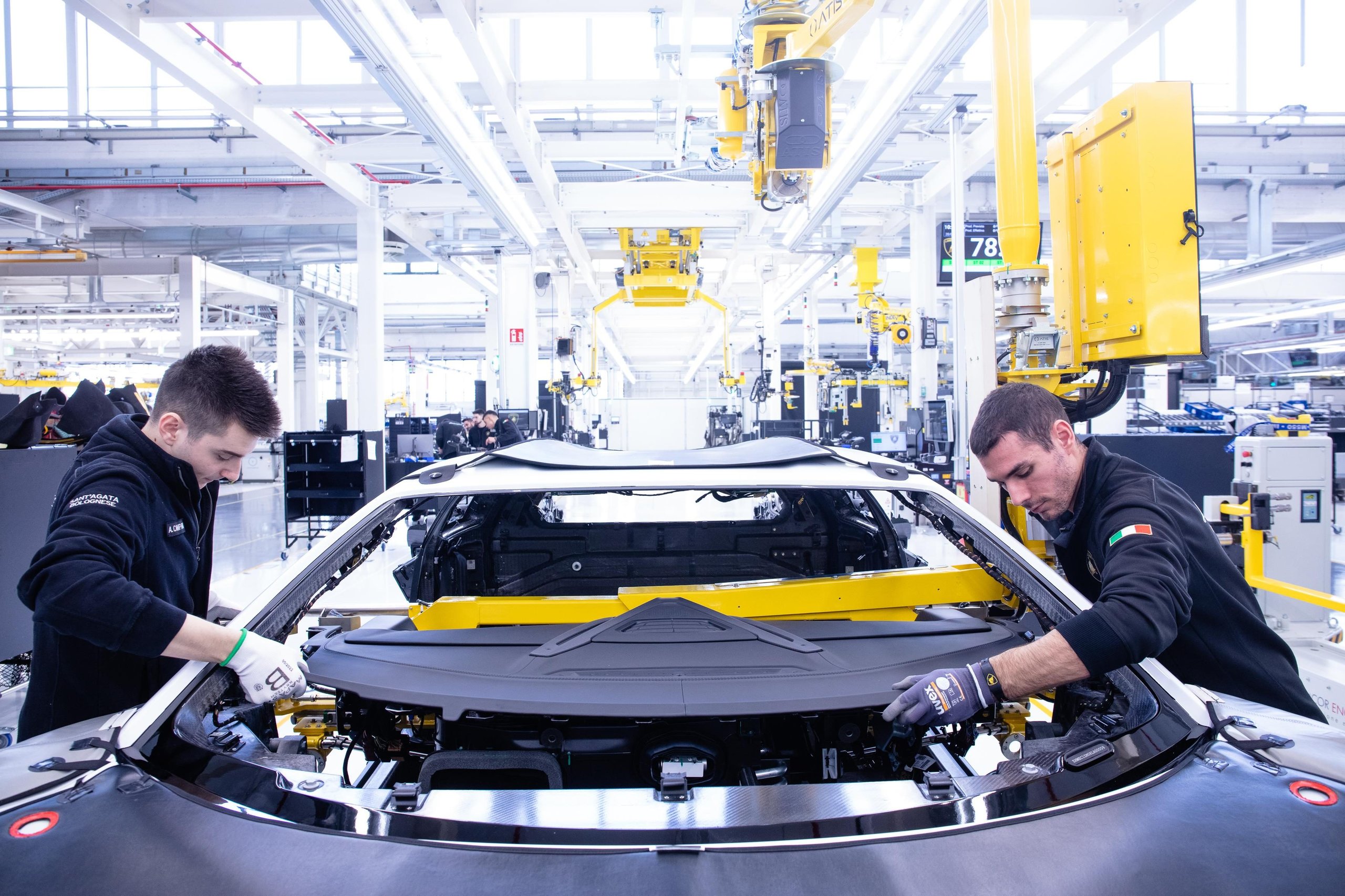 500 Humans, A Fleet Of Cobots, And One Carbon Fiber Tub: How Lamborghini Actually Builds The Revuelto 3 Two lamborghini workers assembling the front section of a white revuelto alongside yellow robotic arms