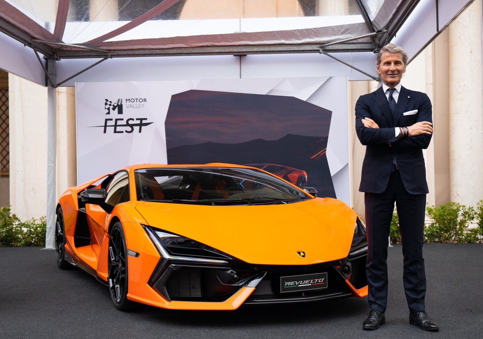 Stephan Winkelmann, Chairman and CEO of Automobili Lamborghini, standing beside an orange Lamborghini Revuelto at Motor Valley Fest 2023 in Modena