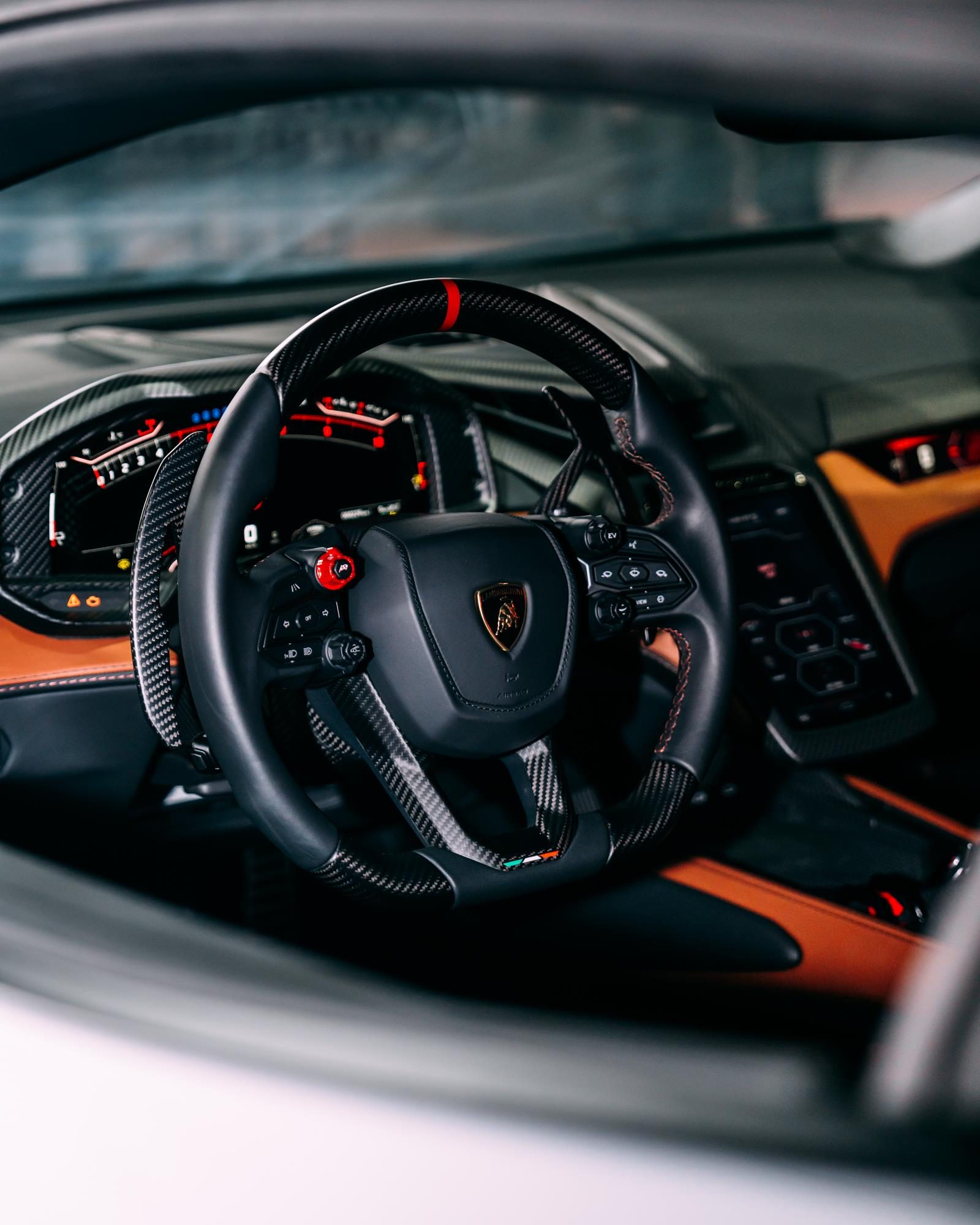 The Lamborghini Revuelto Is The V12 Hybrid That Replaced The Mighty Aventador 5 Lamborghini revuelto interior showing carbon fiber steering wheel, digital instrument cluster, and orange leather dashboard