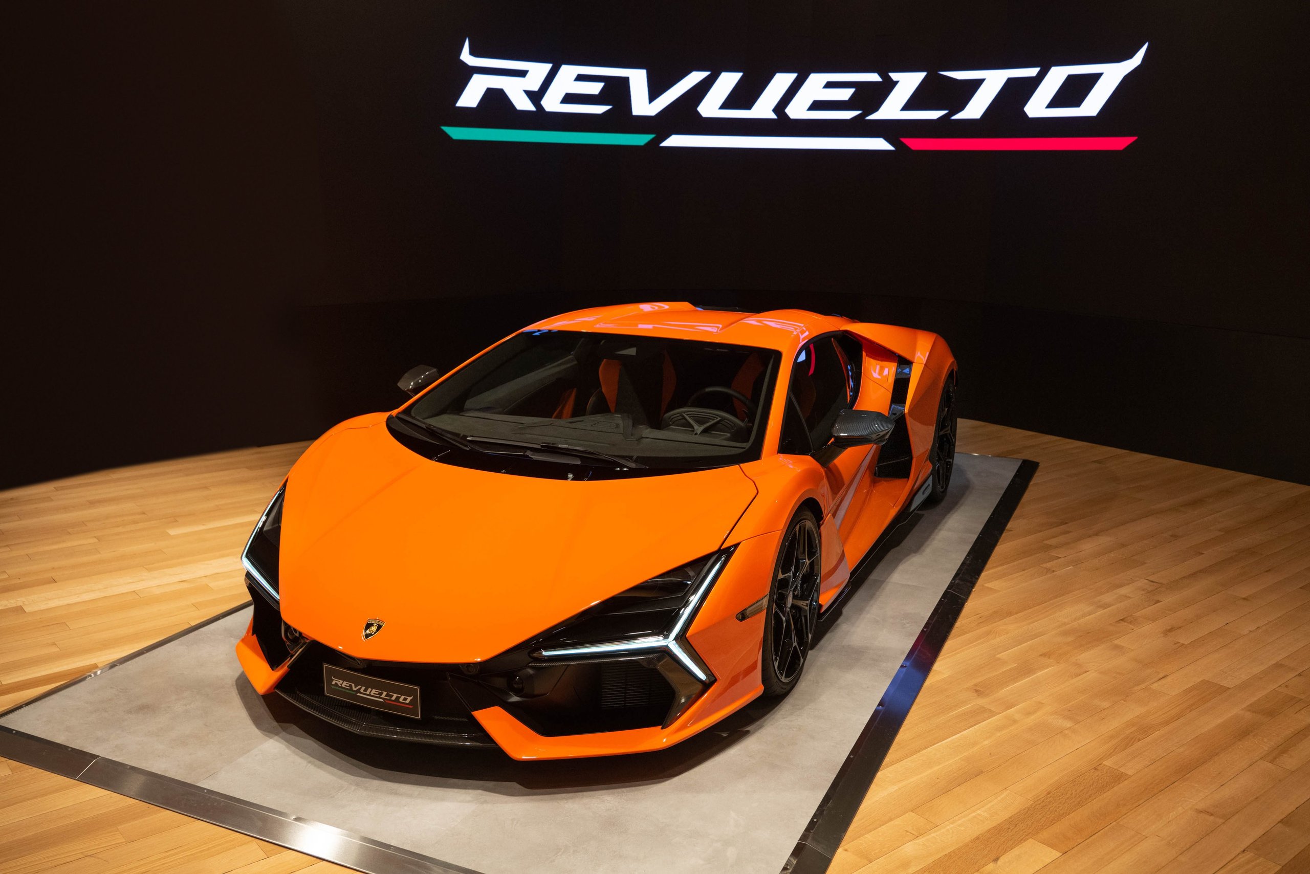 The orange lamborghini revuelto on a raised platform with the illuminated revuelto logo in the background