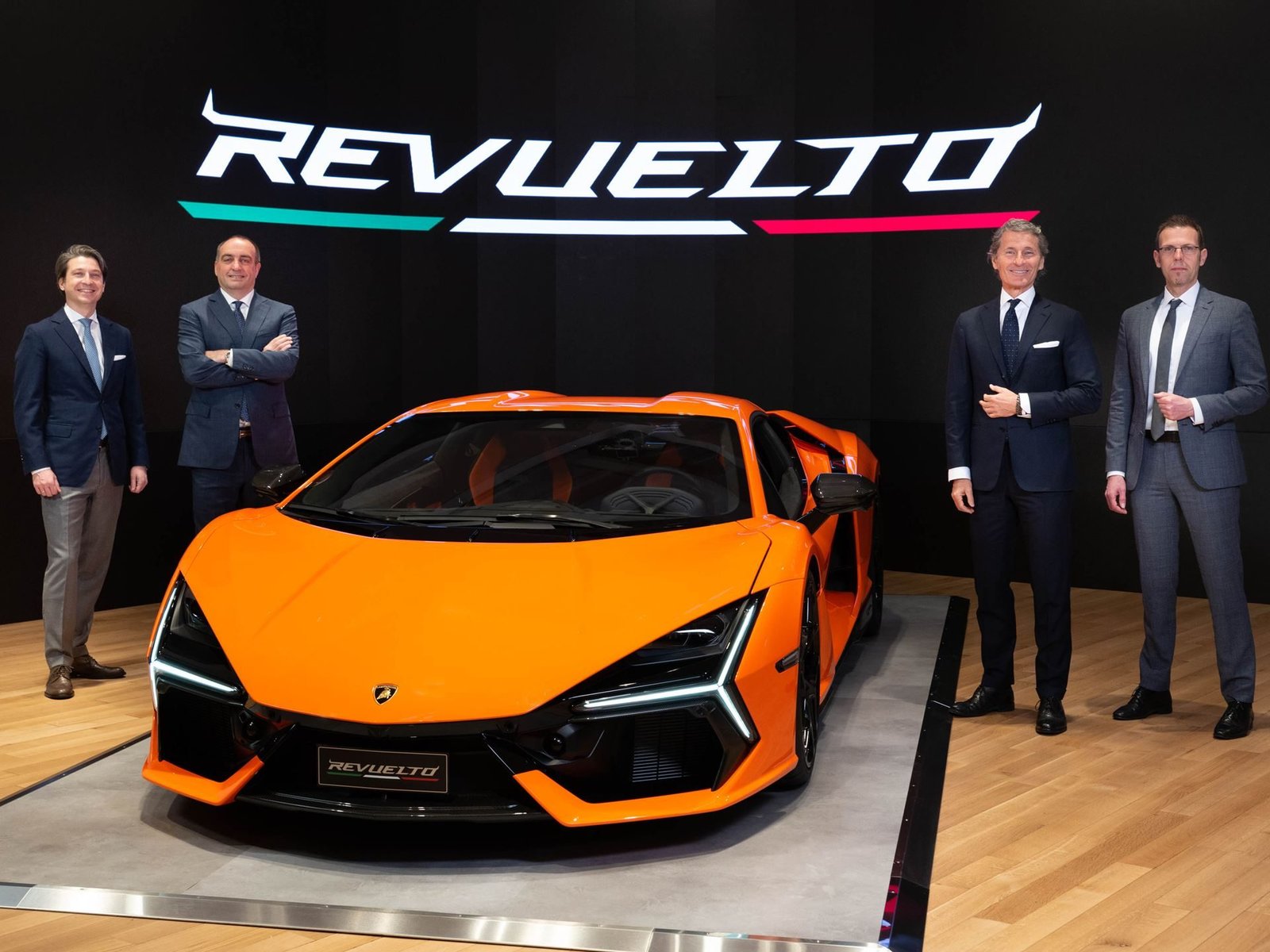 Four Lamborghini executives standing with the orange Revuelto at its US premiere, with the illuminated REVUELTO logo behind them
