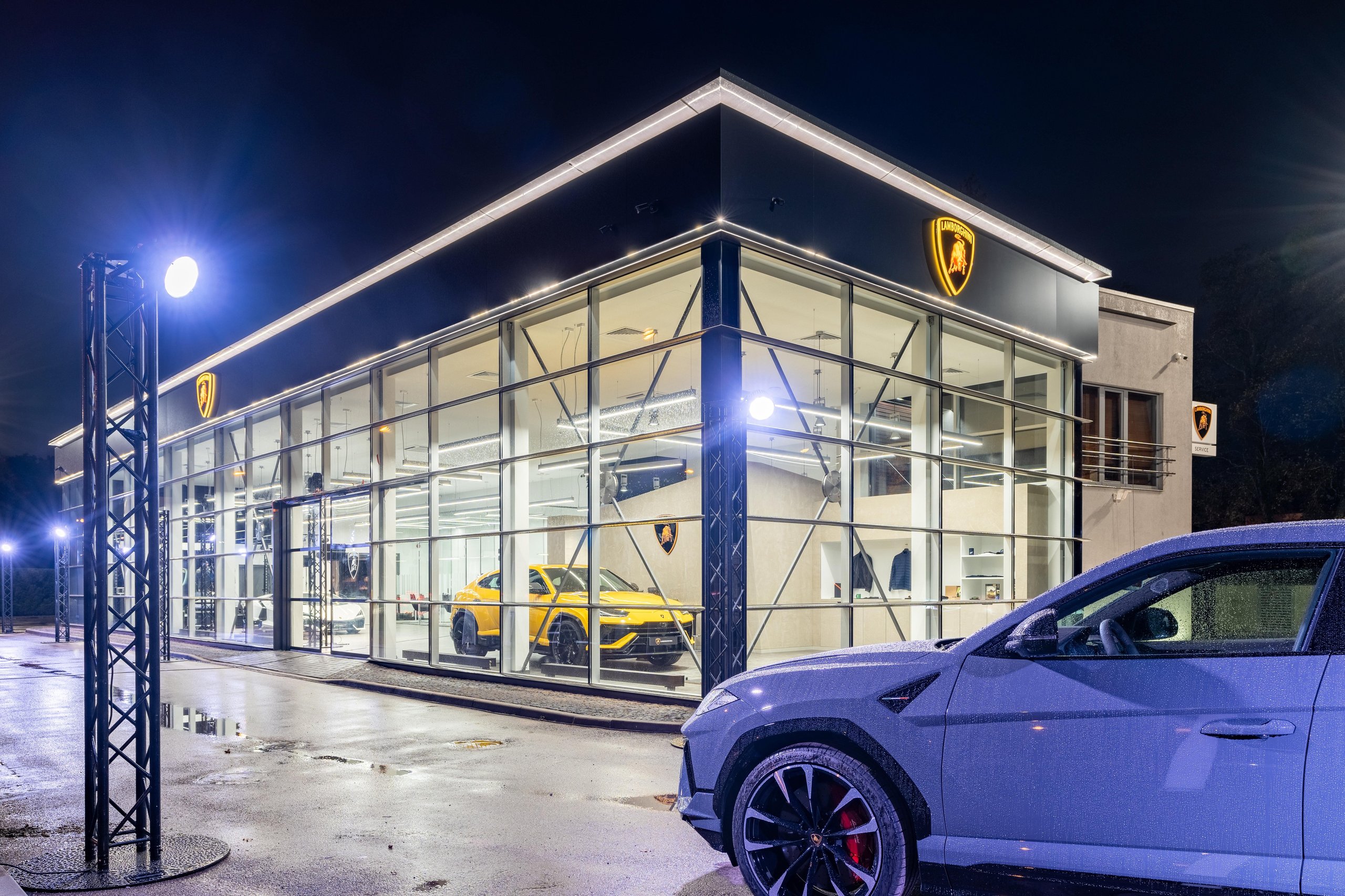 Lamborghini Riga: Why The Brand'S First Latvian Showroom Doubles As A Hybrid-Era Statement 5 The lamborghini riga dealership illuminated at night with a grey urus parked outside and a yellow urus visible through large glass windows