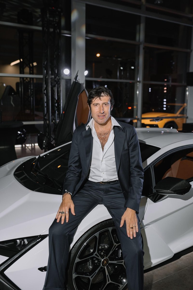A man in a dark suit sits casually on the hood of a white Lamborghini Revuelto with an open scissor door visible behind him at the Riga showroom opening