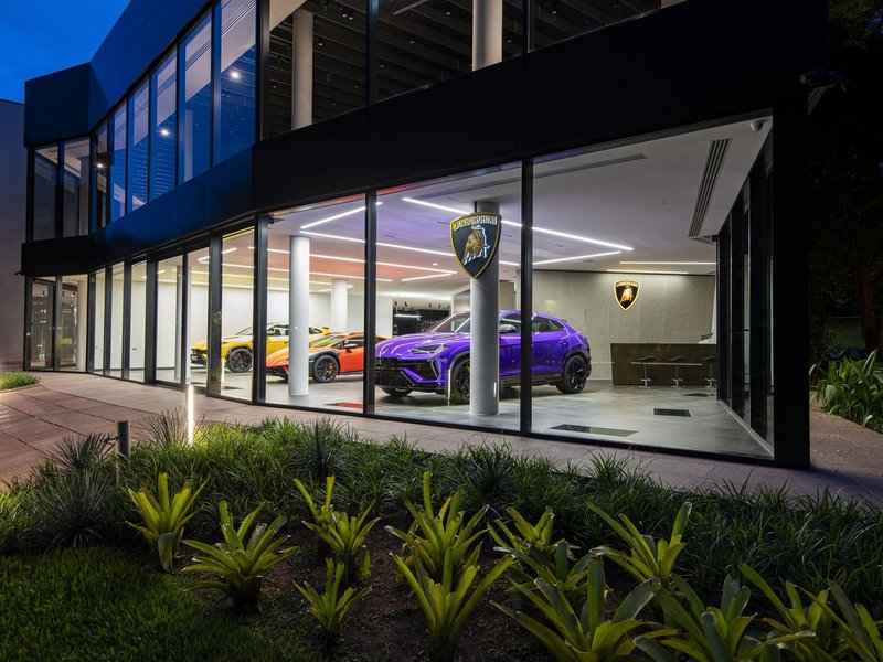 Evening exterior of the new Lamborghini São Paulo showroom with a purple Urus, orange Huracán Sterrato, and yellow Urus visible through floor-to-ceiling glass windows